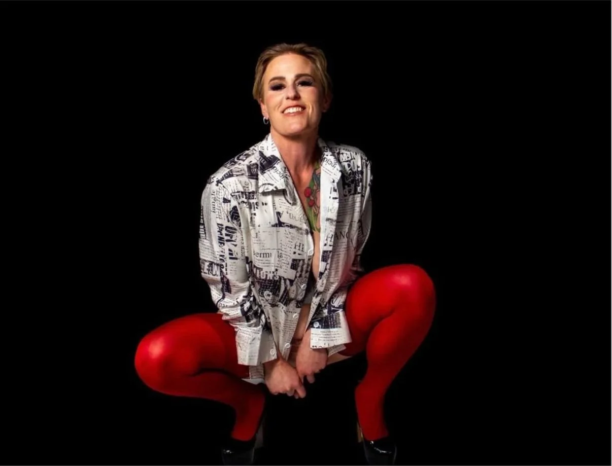 A woman with short blonde hair, wearing a newspaper-print shirt, red tights, and high heels, squatting with legs apart against a black background.