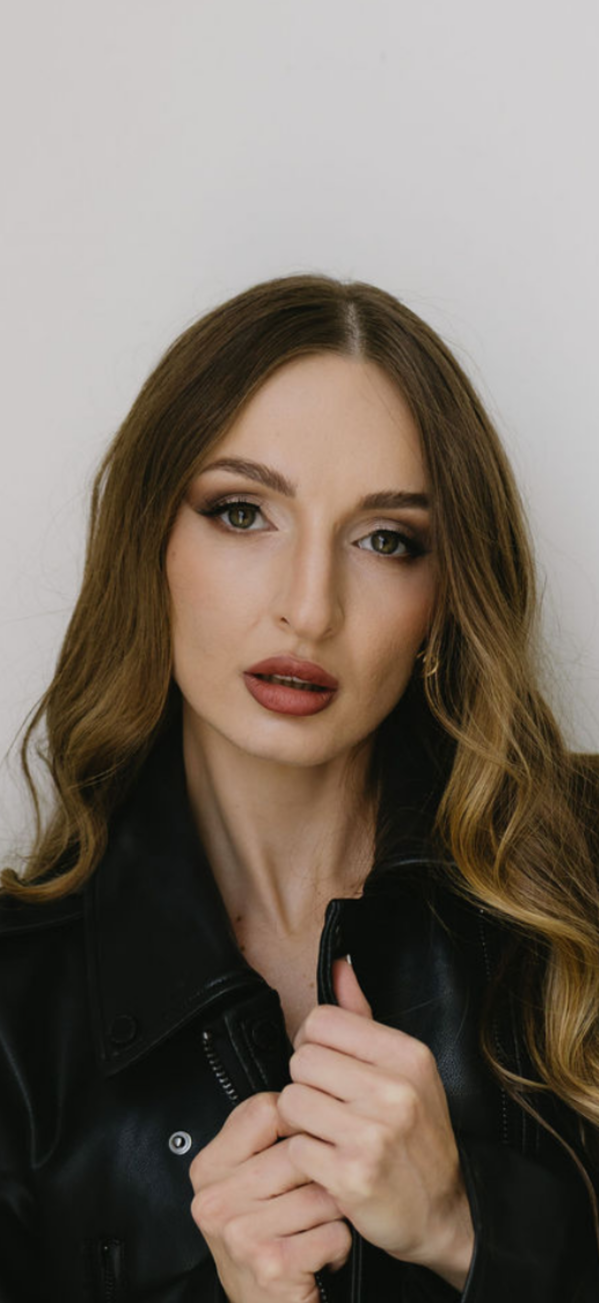 A portrait of a woman with long wavy brown hair, wearing makeup, and a black leather jacket, holding the collar with an intense expression.