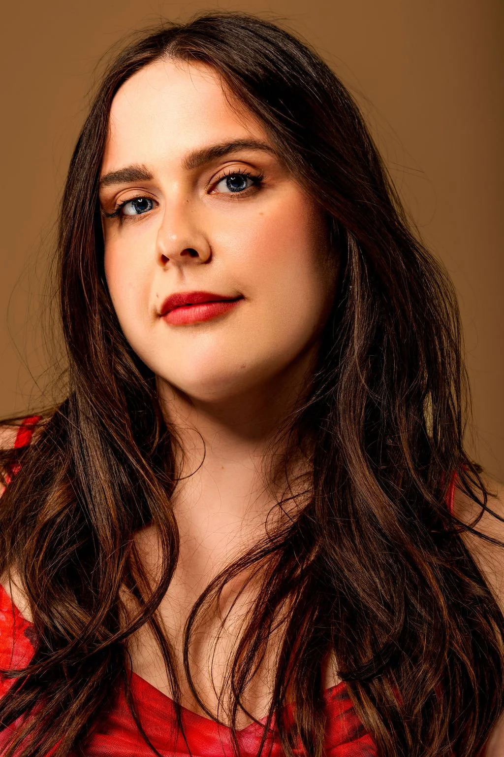 A woman with long brown hair, blue eyes, and red lipstick, wearing a red top, posing against a brown background.