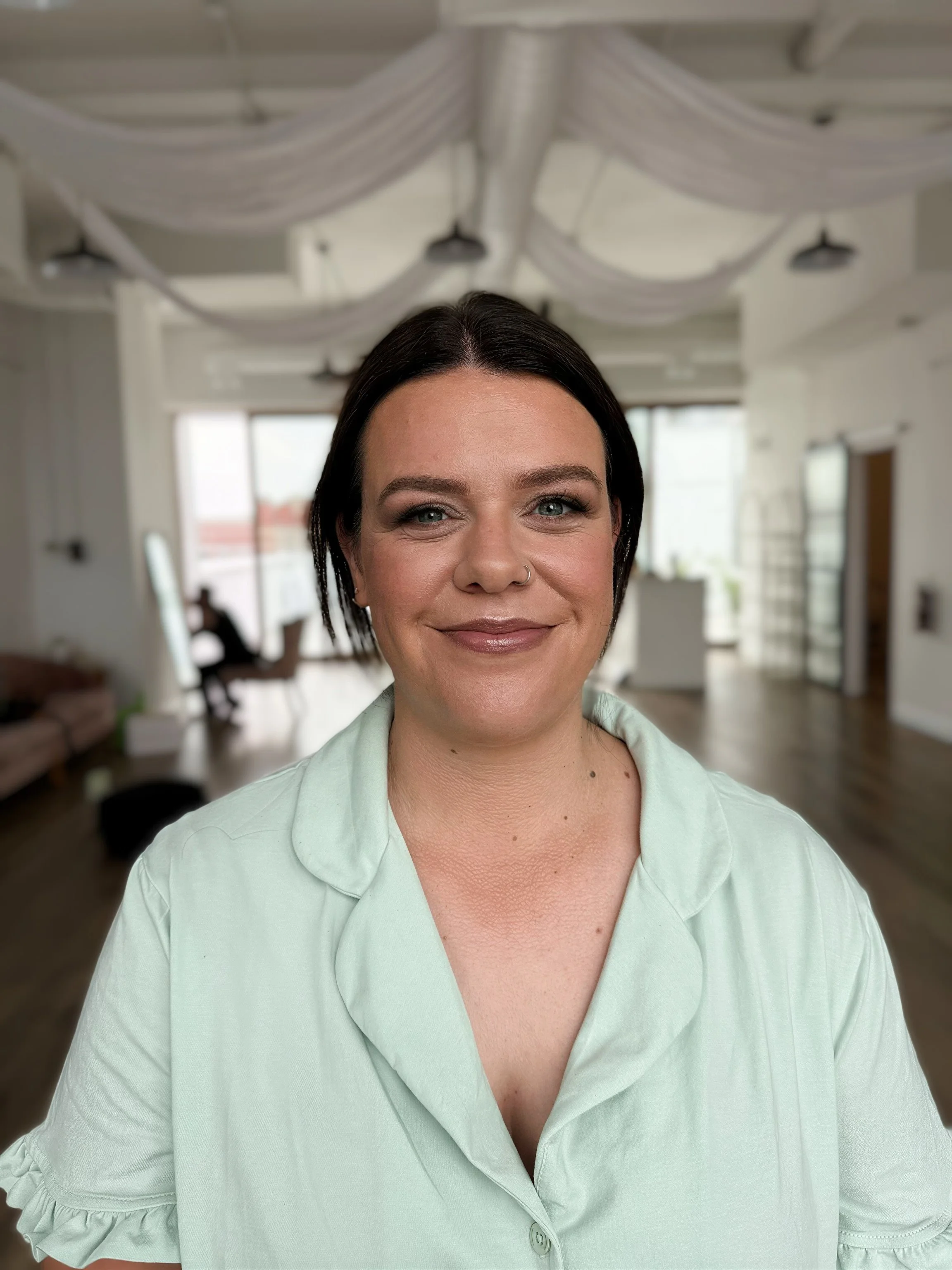 A woman with dark hair and light skin smiling at the camera in a bright, modern indoor space with large windows.