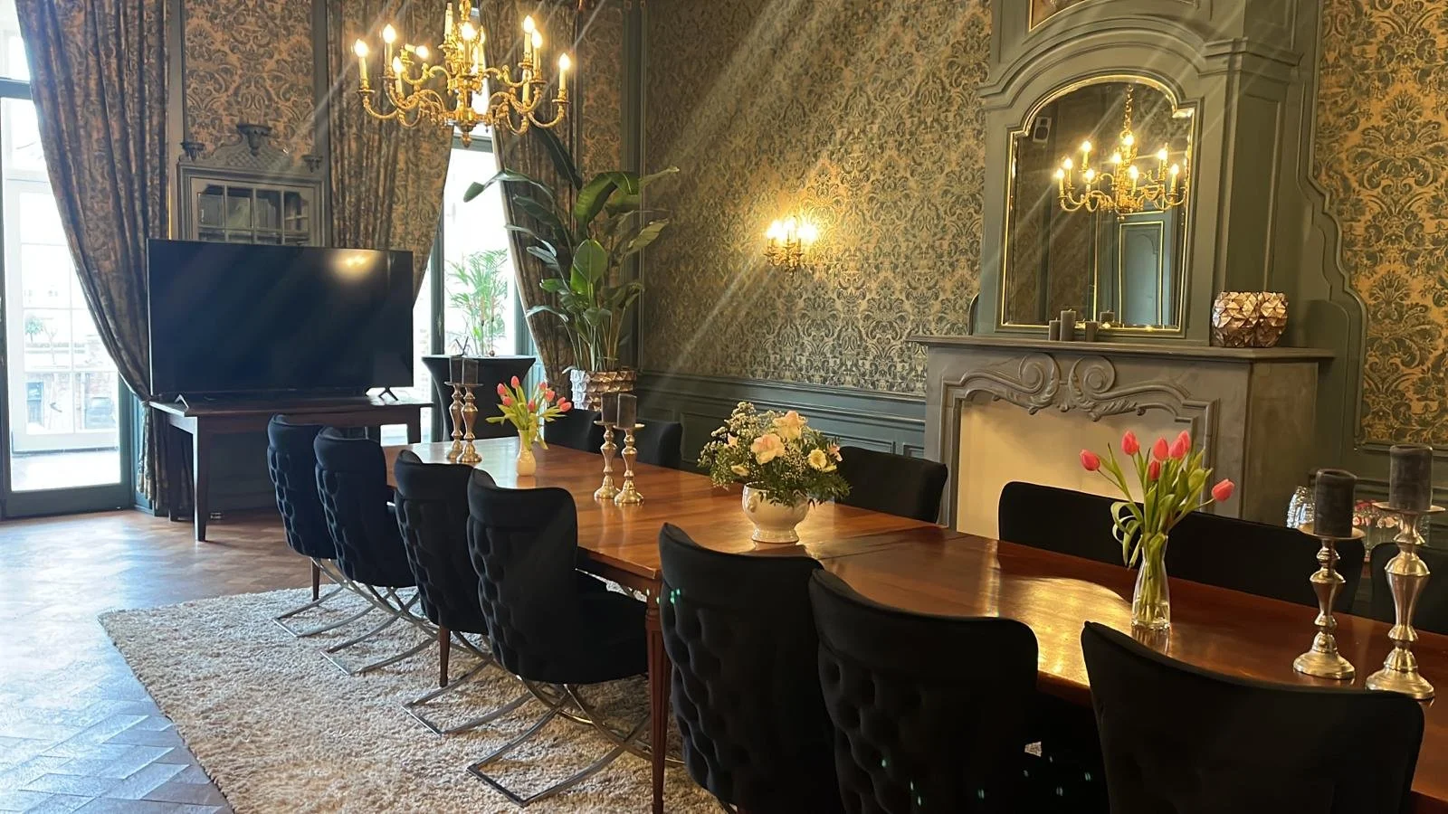 Een elegante eetkamer met een grote houten tafel, zwarte stoelen, bloemen in vazen, een grote tv, kroonluchters, grote ramen met gordijnen en een decoratieve schouw met spiegel.