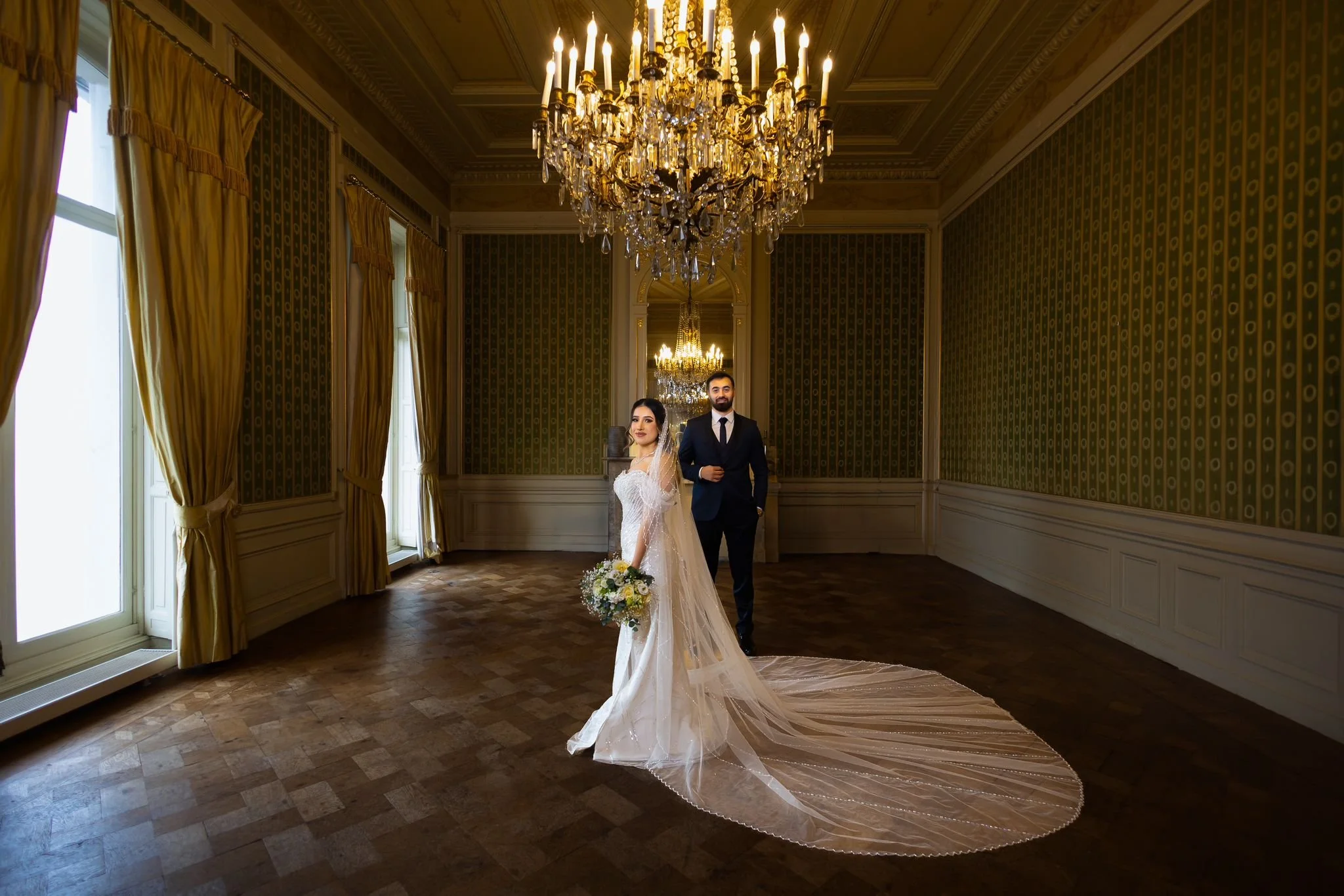 Bruide en bruidegom in een elegant, grote kamer met gouden details en kristallen kroonluchters, de bruid draagt een wit huwelijkss jurk met een lange strooien rok en houdt een boeket bloemen, de bruidegom draagt een zwart pak en witte blouse.