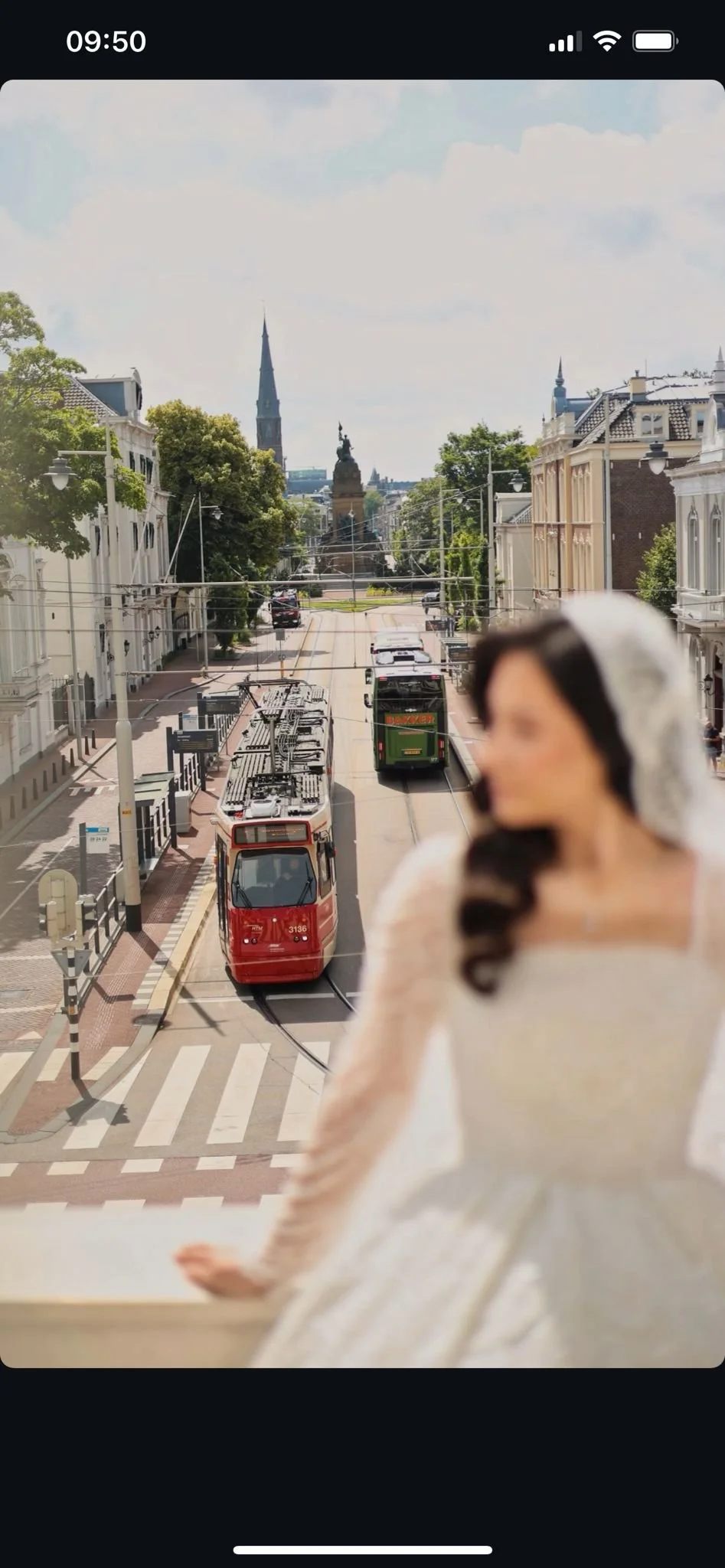 Een vrouw in een feestelijke witte jurk en haarcup met zwart haar, op de achtergrond een straat met trams, historische gebouwen en een standbeeld, vermoedelijk in een Europese stad.