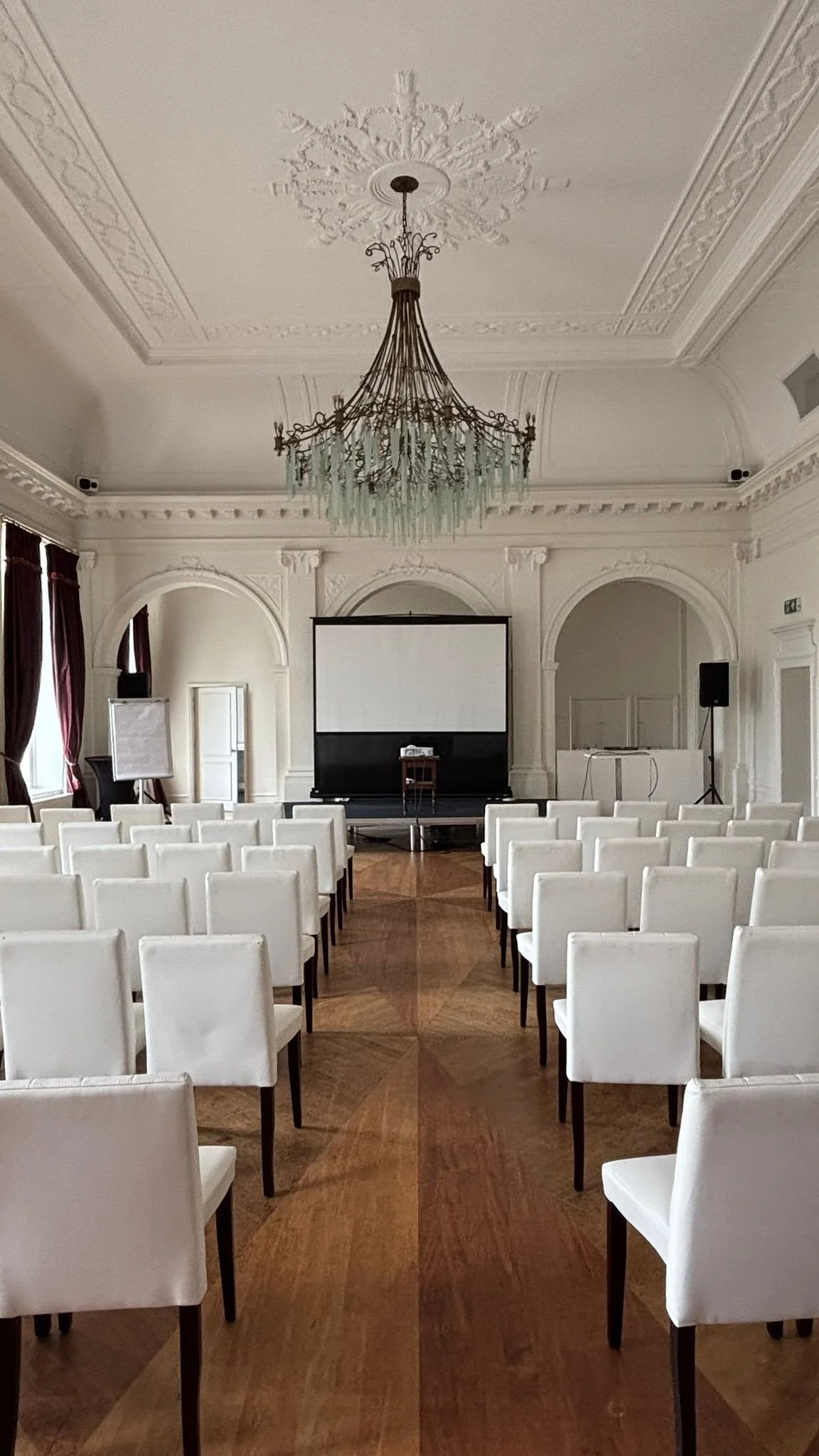 Een stijlvolle vergaderzaal met wit gestoffeerde stoelen, een groot scherm en een chandelier in een elegante witte kamer.