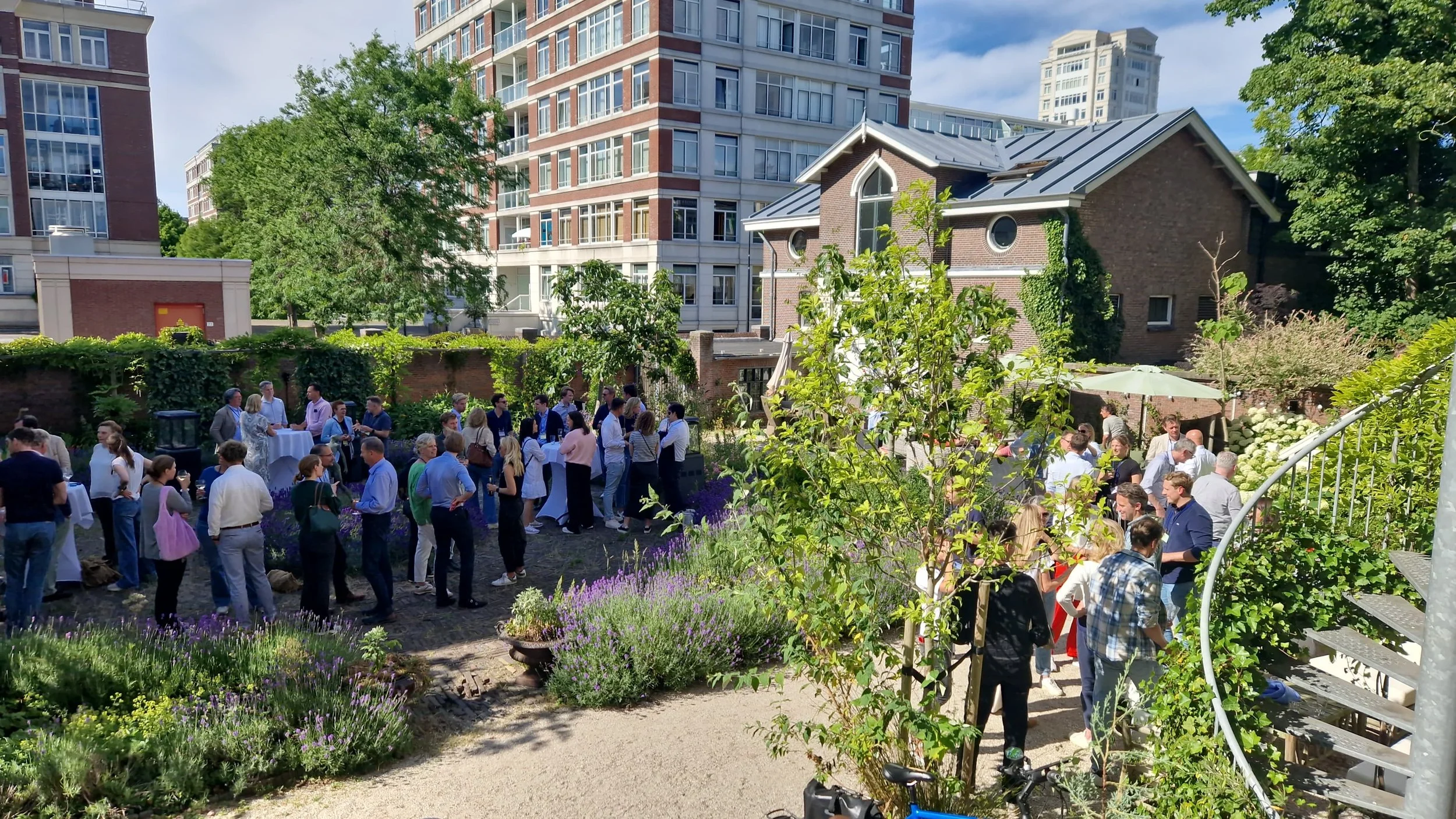 Bijeenkomst in een tuin met veel mensen, bloemen en moderne gebouwen op de achtergrond, tijdens een zonnige dag.