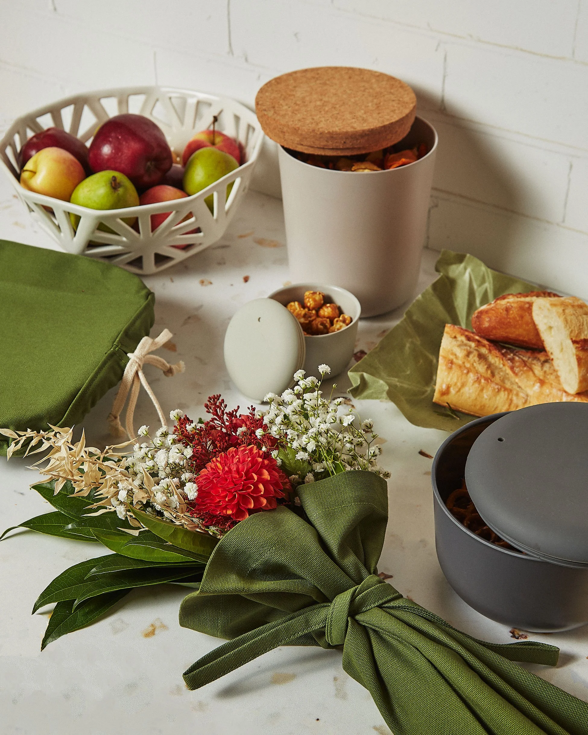 Lifestyle photography image of a table with a bouquet of flowers, a green bag, a basket of apples, a container of cereal, a jar of caramel popcorn, a loaf of bread, and a gray container with a lid.