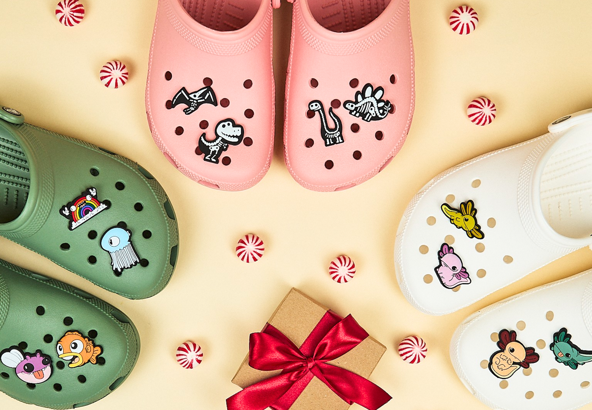 Ecommerce Audit Services - Colorful Crocs in pink, green, white, and cream with cartoon dinosaur and animal charms, arranged in a circular pattern with peppermint candies and a gift box with red ribbon in the center, on a yellow background.