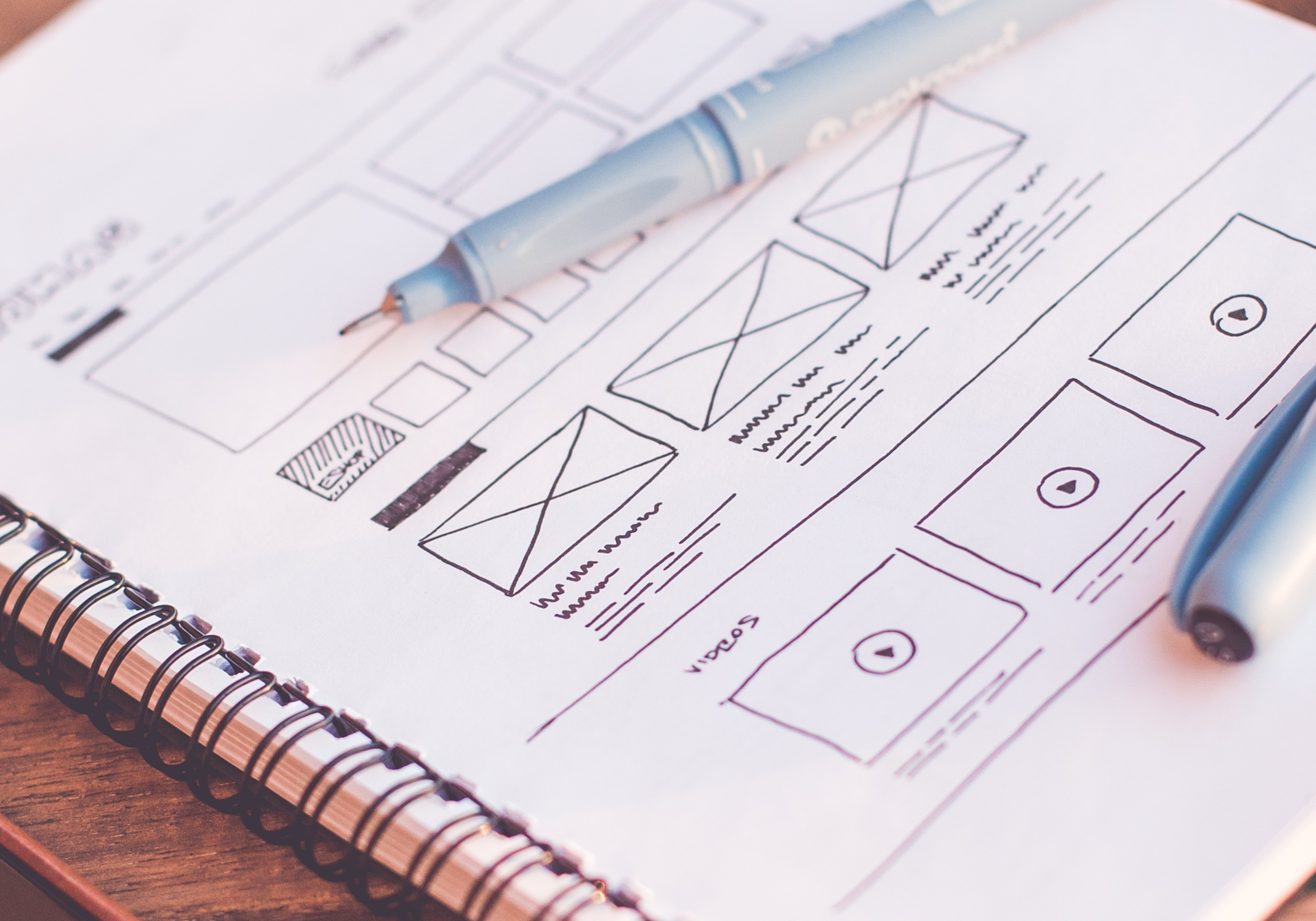 A hand-drawn wireframe sketch of a website or app layout on a spiral notebook page with a blue marker marker resting on it.