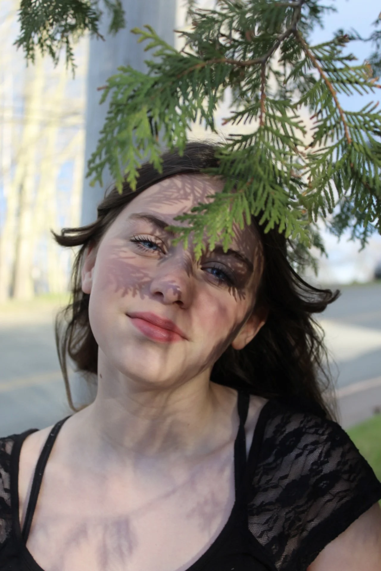 A young woman with light skin and dark hair standing outdoors near a tree with green leaves, which cast shadow patterns on her face. The background shows a blurred street and trees, suggesting a park or neighborhood setting.