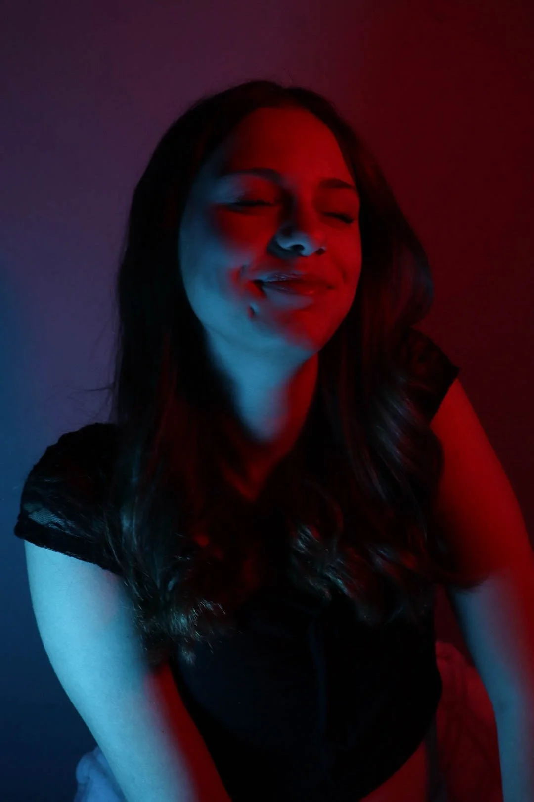 A woman with long wavy hair is smiling with her eyes closed, illuminated by red and blue lighting, wearing a black top.