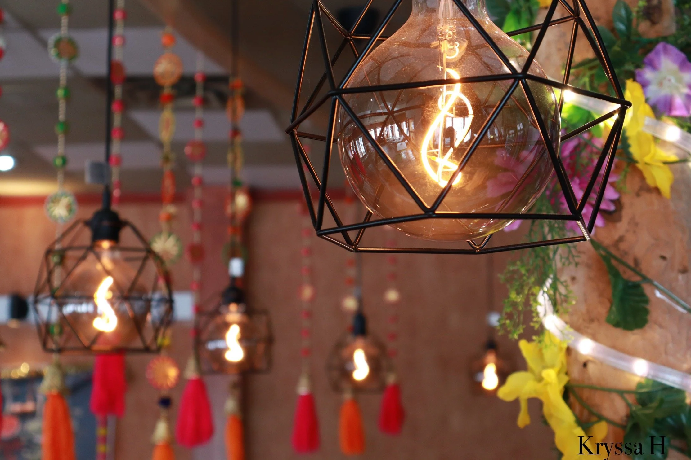 Decorative hanging light fixtures with exposed bulbs in geometric metal frames, with colorful hanging ornaments and flowers in the background. photo taken in cape breton.