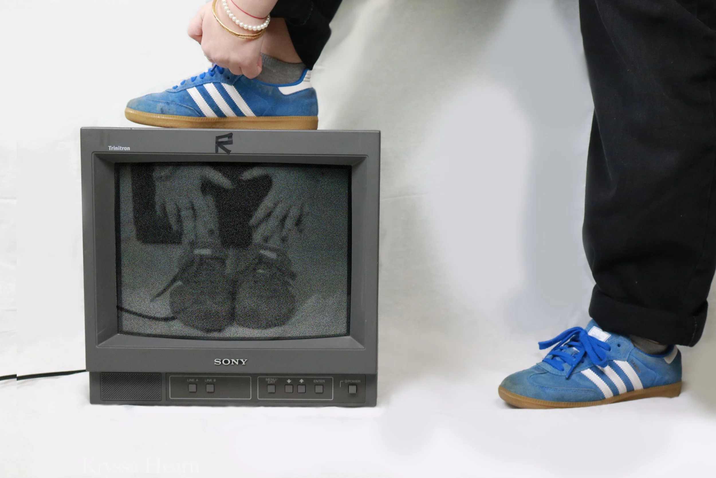 Person wearing blue sneakers with white stripes stepping on an old Sony television set, which displays a reflection of feet and hands on a gray background.