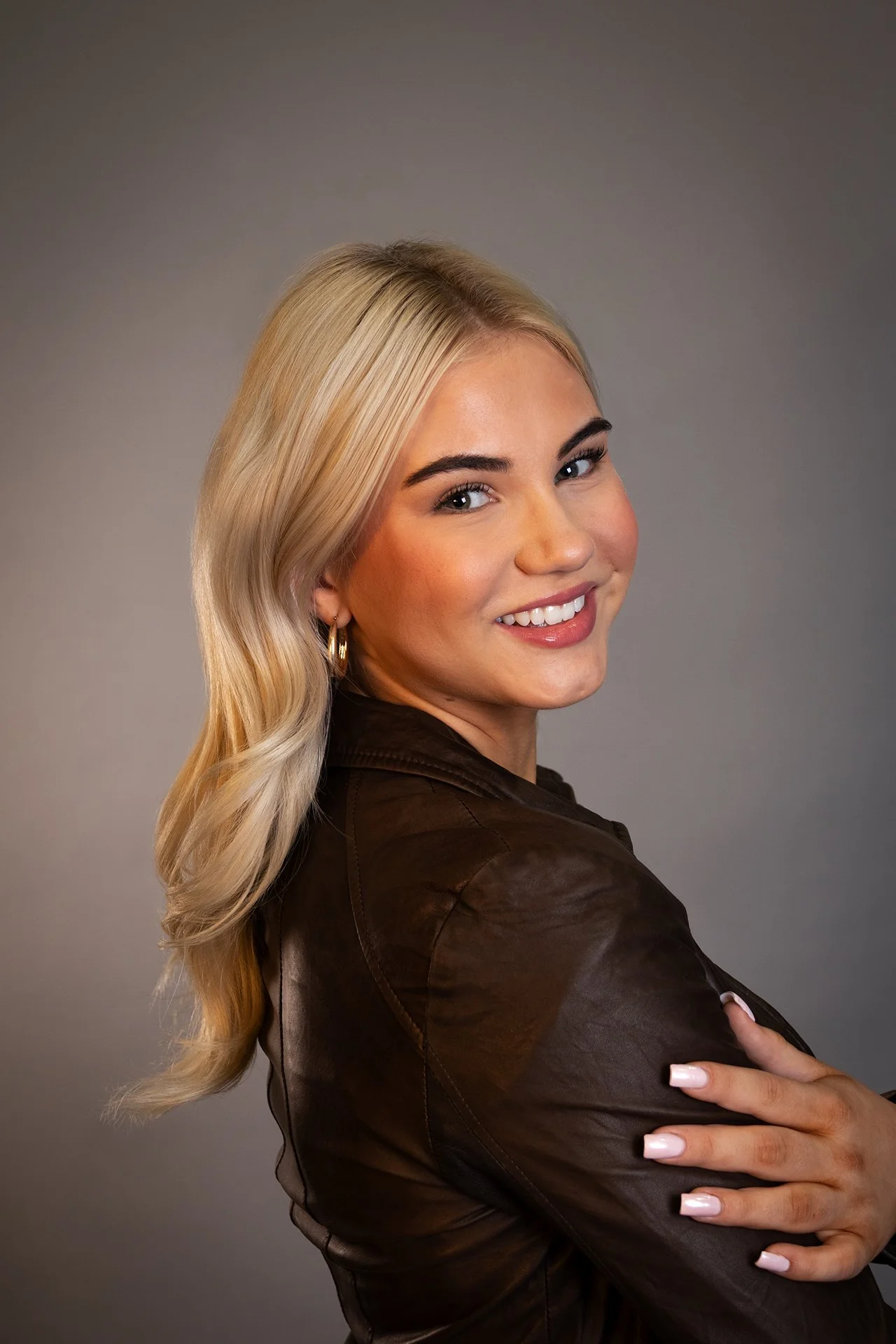 A smiling woman with long blonde hair, wearing a brown leather jacket and earrings, posing with her arms crossed against a gray background.