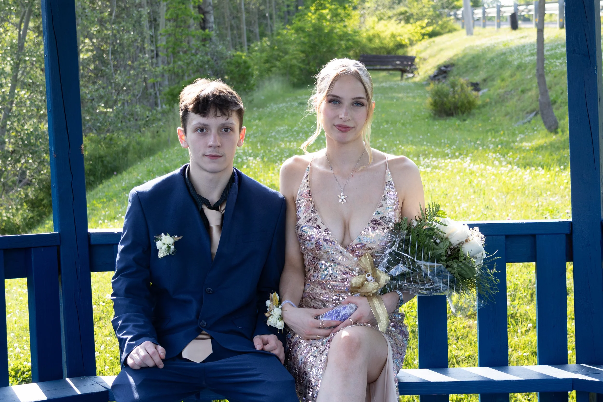 A young man and woman sitting side by side on a blue wooden bench outdoors, with greenery and trees in the background. The woman holds a bouquet of flowers and wears a sparkly dress, while the man is dressed in a dark suit.
