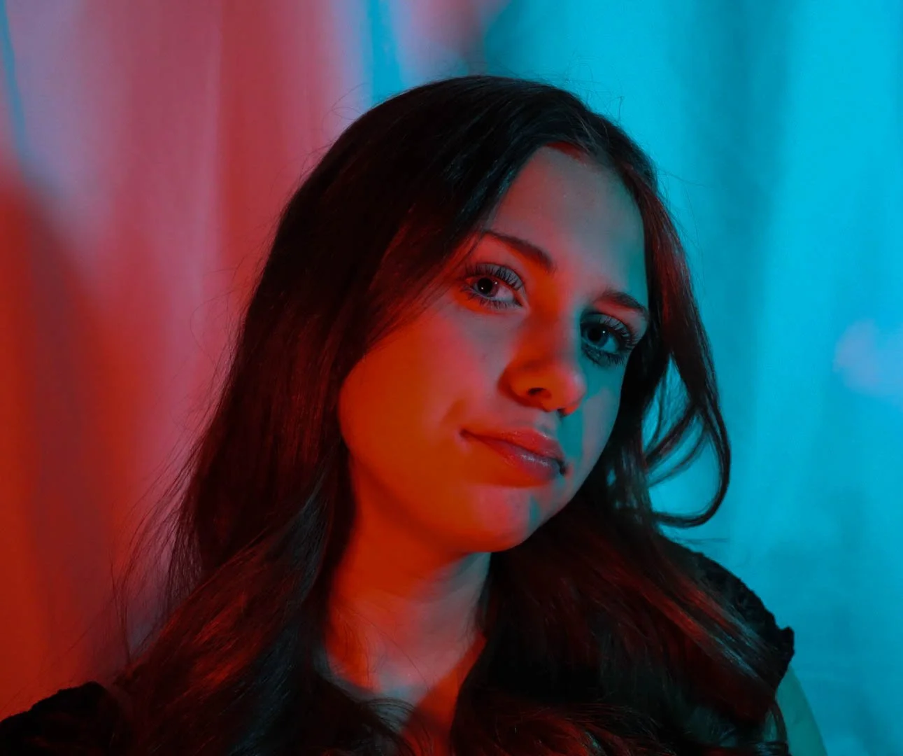 Portrait of a young woman with long dark hair, illuminated by red and blue lighting, smiling slightly.