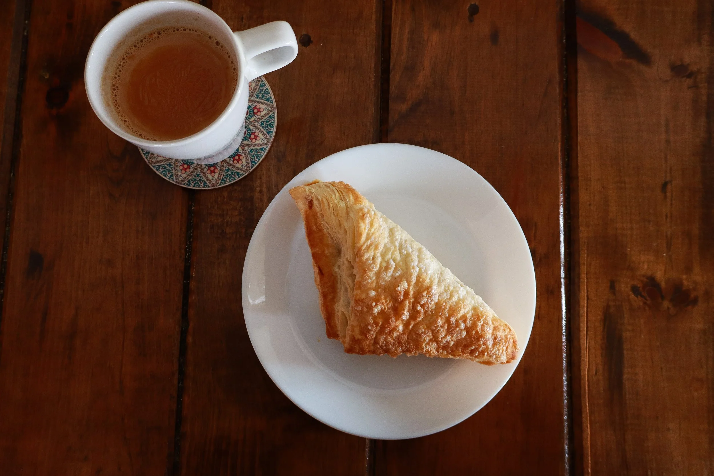 A white plate with a triangular piece of fried pastry, a white mug of tea on a decorative coaster, on a wooden table.