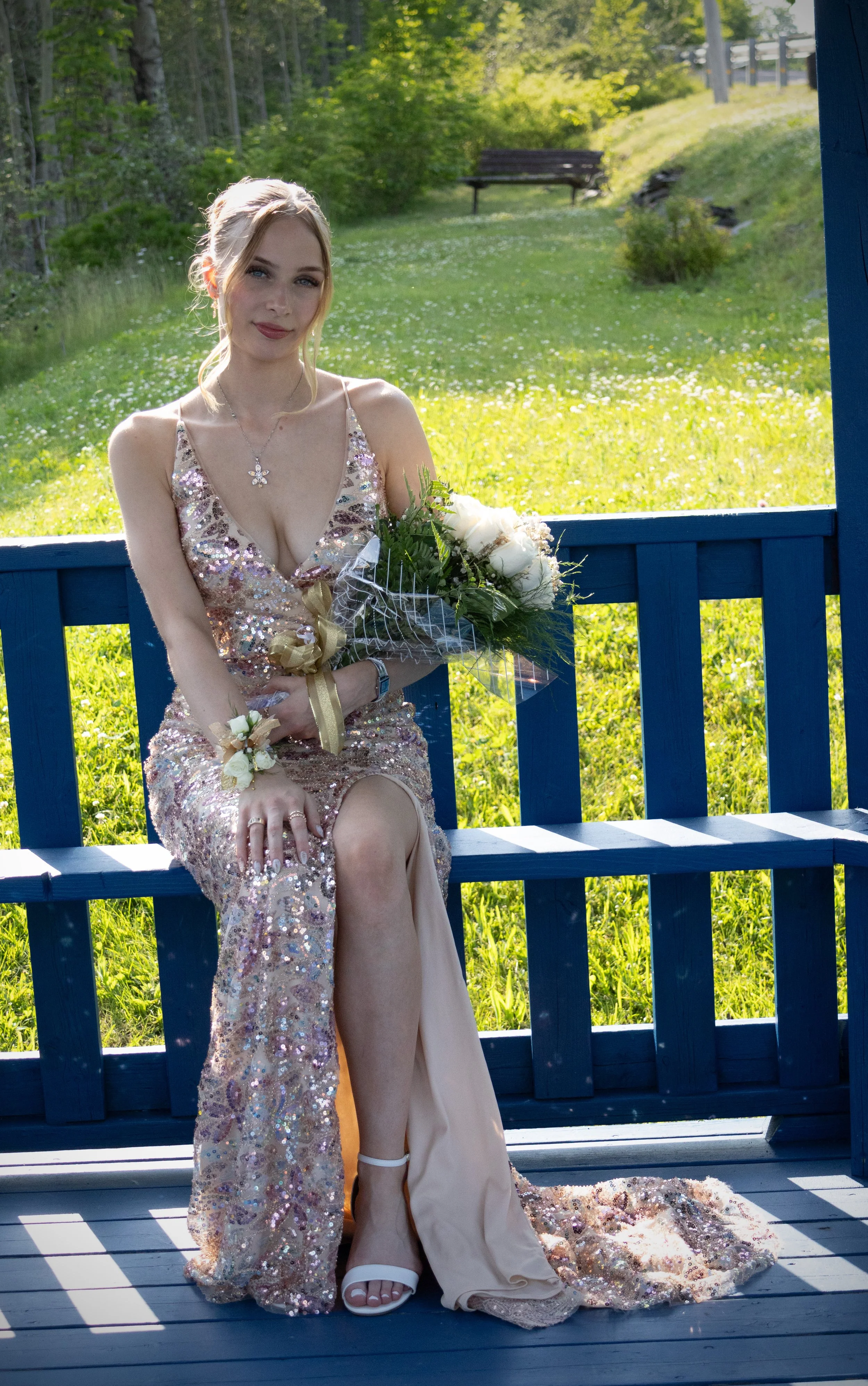 A young woman in a sparkly, sleeveless gown sits on a blue wooden bench outdoors, holding a bouquet of flowers and wearing a corsage and a flower necklace. She has blonde hair styled in an updo and is looking at the camera with a slight smile, with g