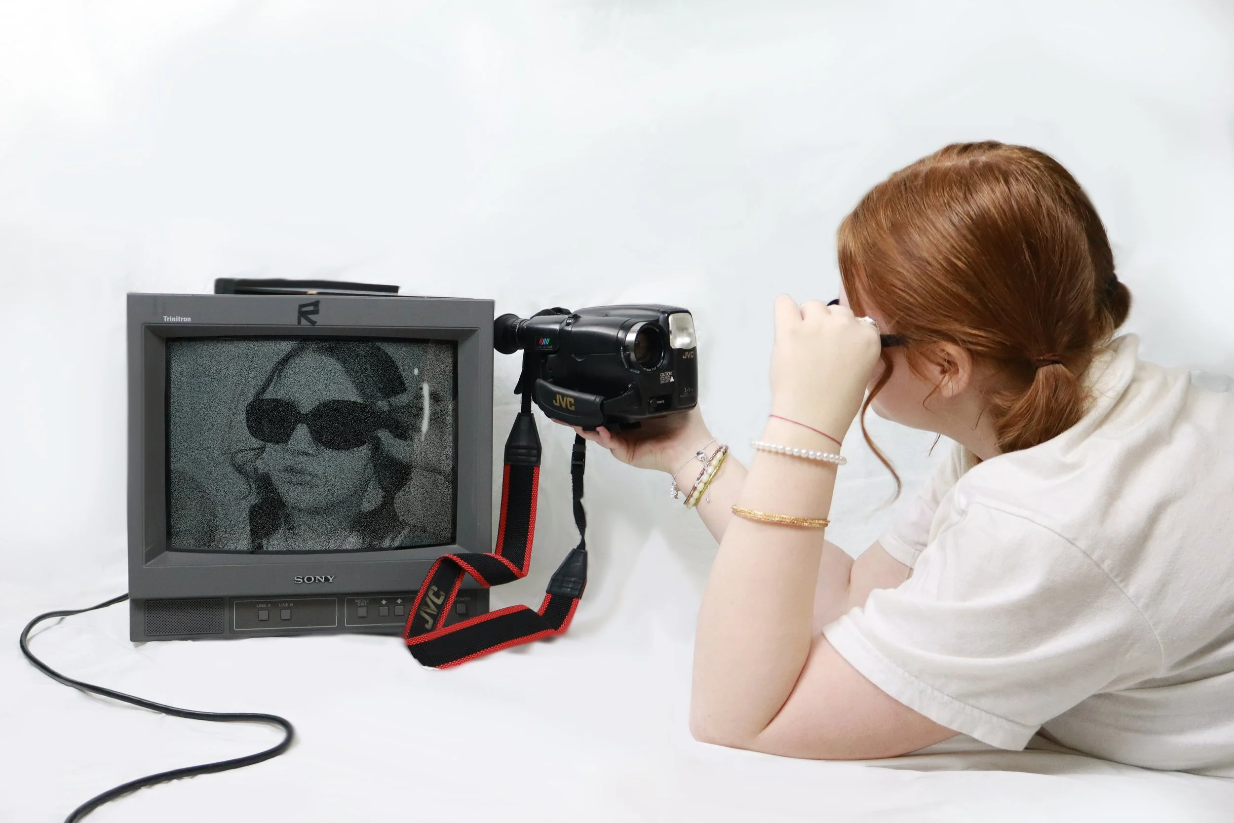 A woman with red hair and glasses lying on her side, holding a video camera to her eye, filming a television screen displaying a black-and-white image of a woman wearing sunglasses.