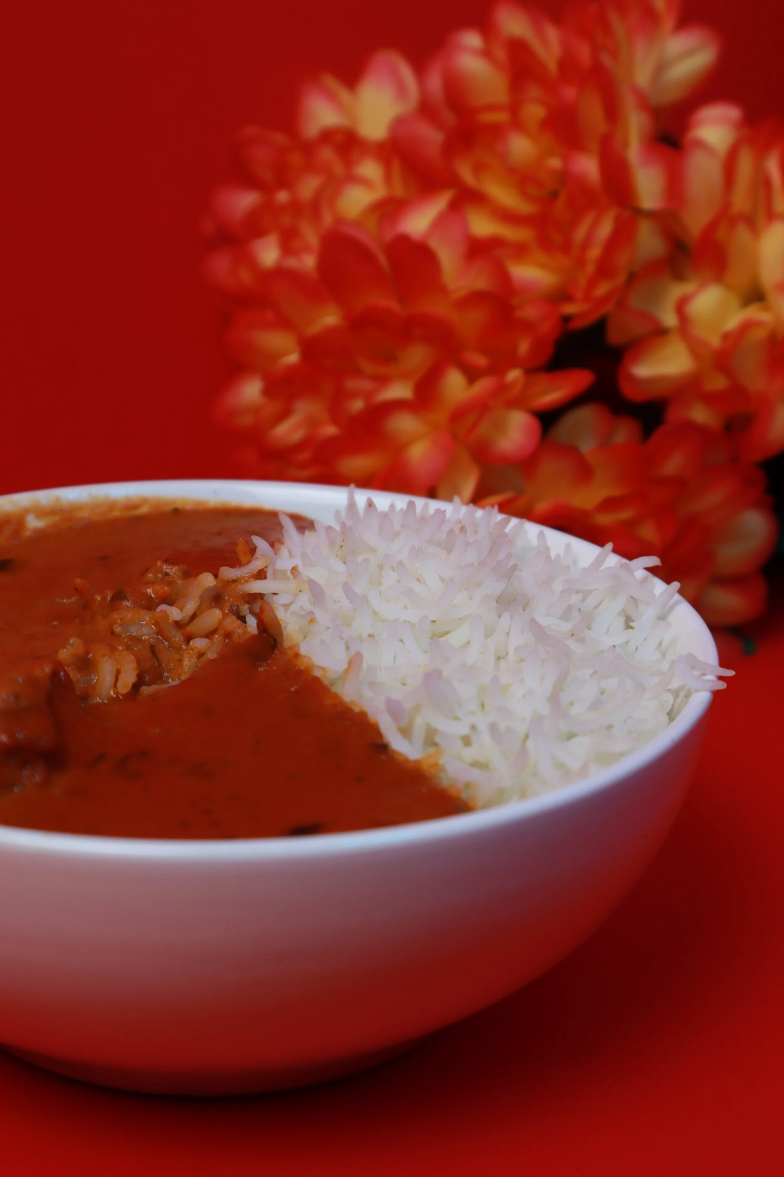 A white bowl of rice and beef stew on a red surface with orange flowers in the background.