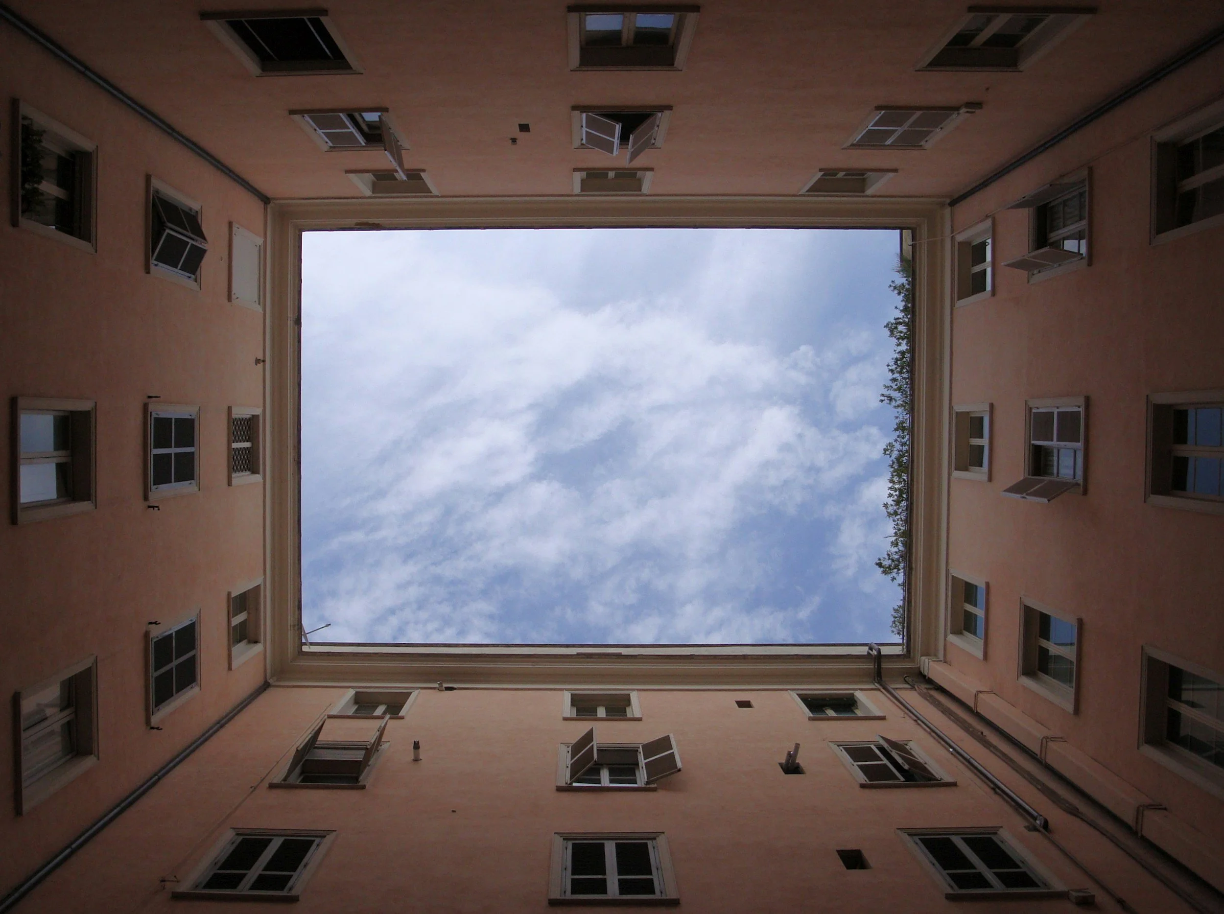 Looking up from the courtyard of a residential building shows a blue sky with scattered clouds through the open roof square, surrounded by beige walls with multiple windows slightly open.
