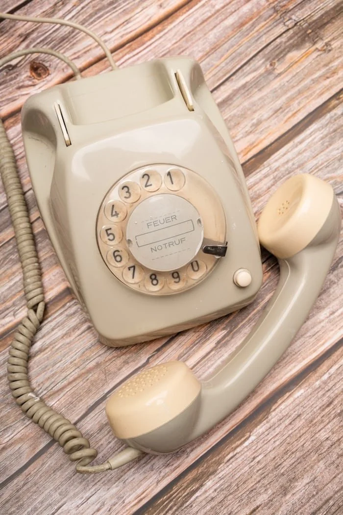An off-white vintage rotary phone with a circular dial and a coiled cord on a wooden surface.