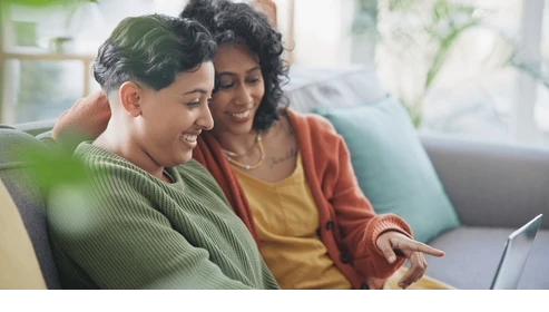 Two women sitting on a couch, looking at a smartphone and smiling in a cozy, well-lit living room.