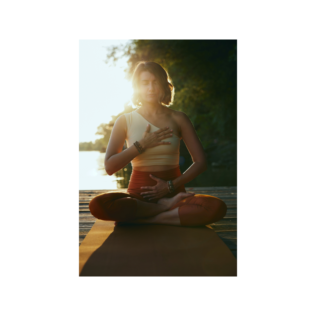 Woman practicing yoga outdoors at sunset, sitting cross-legged on a mat near a body of water with trees in the background.