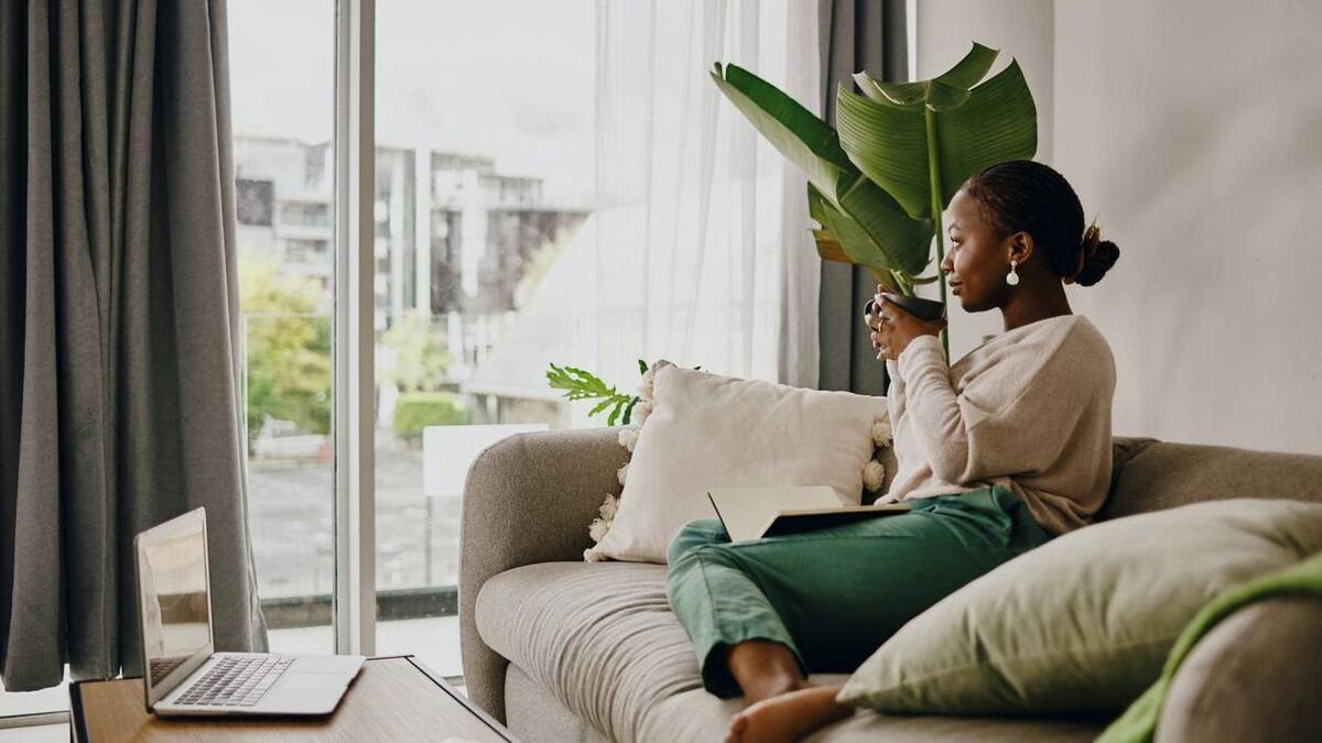A woman sitting on a sofa in a modern living room, holding a large green leafy plant, with a laptop and tablet on the sofa, and large windows with curtains in the background.