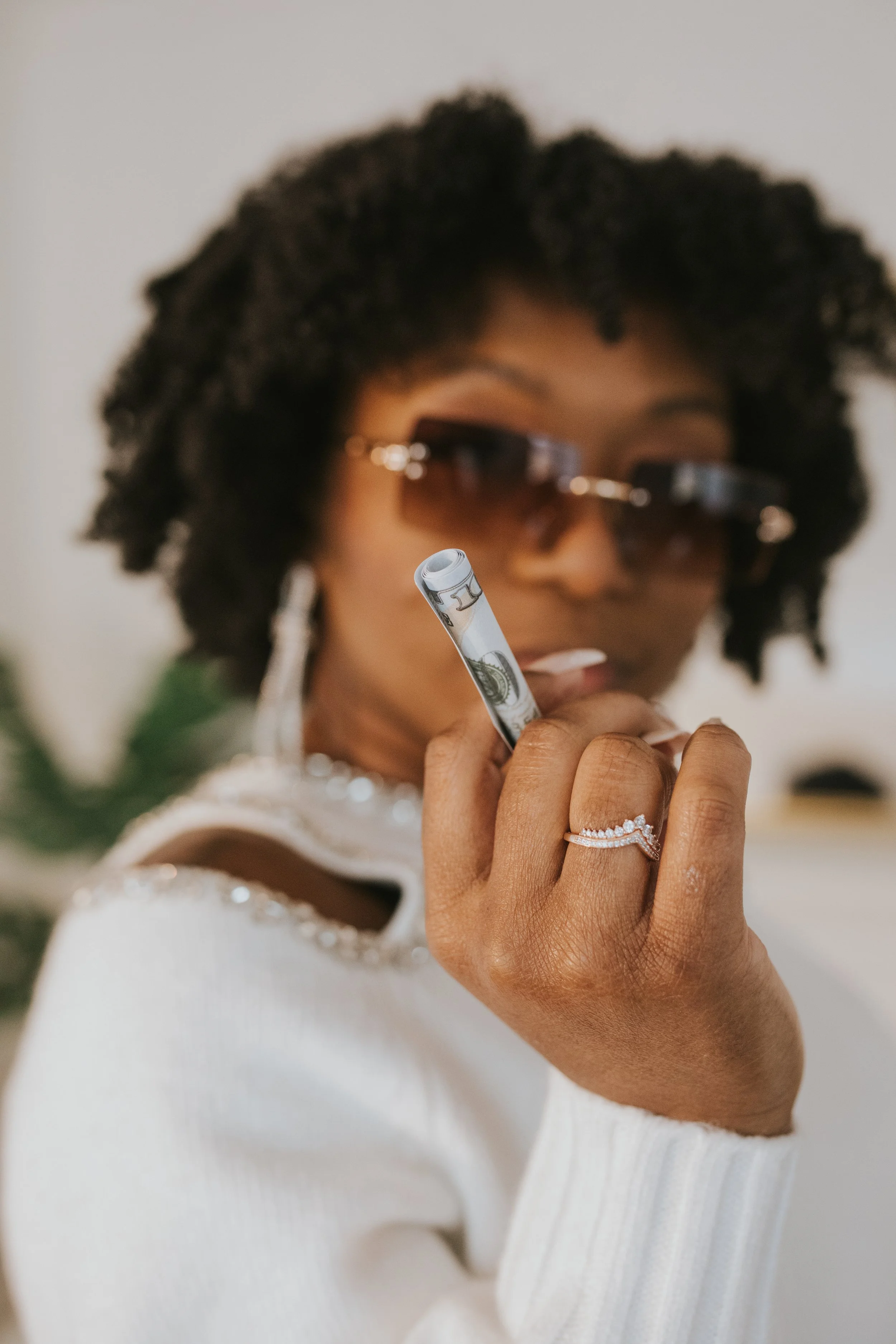 A woman with curly hair and sunglasses holding a rolled hundred-dollar bill.