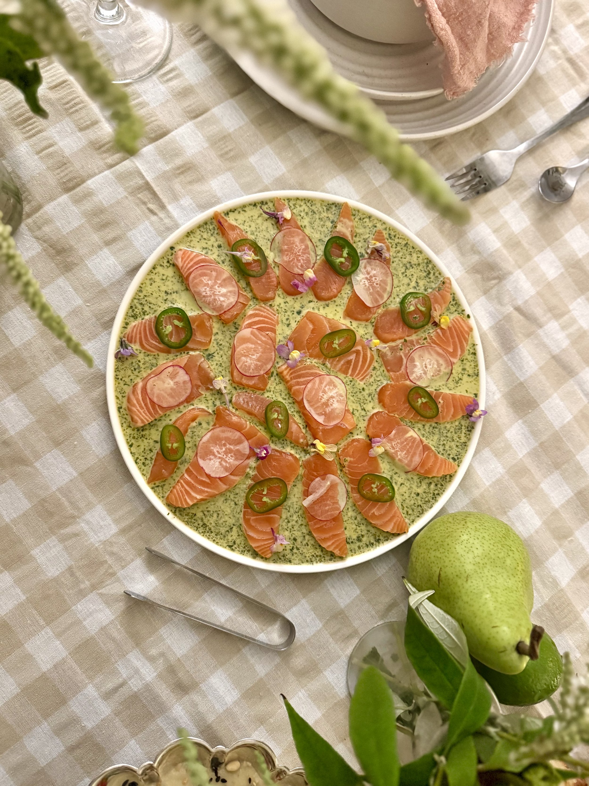 A circular dish with slices of salmon, jalapeño slices, and edible flowers on a green sauce or mousse, garnished with lemon slices and pink edible flower petals, on a checkered tablecloth.