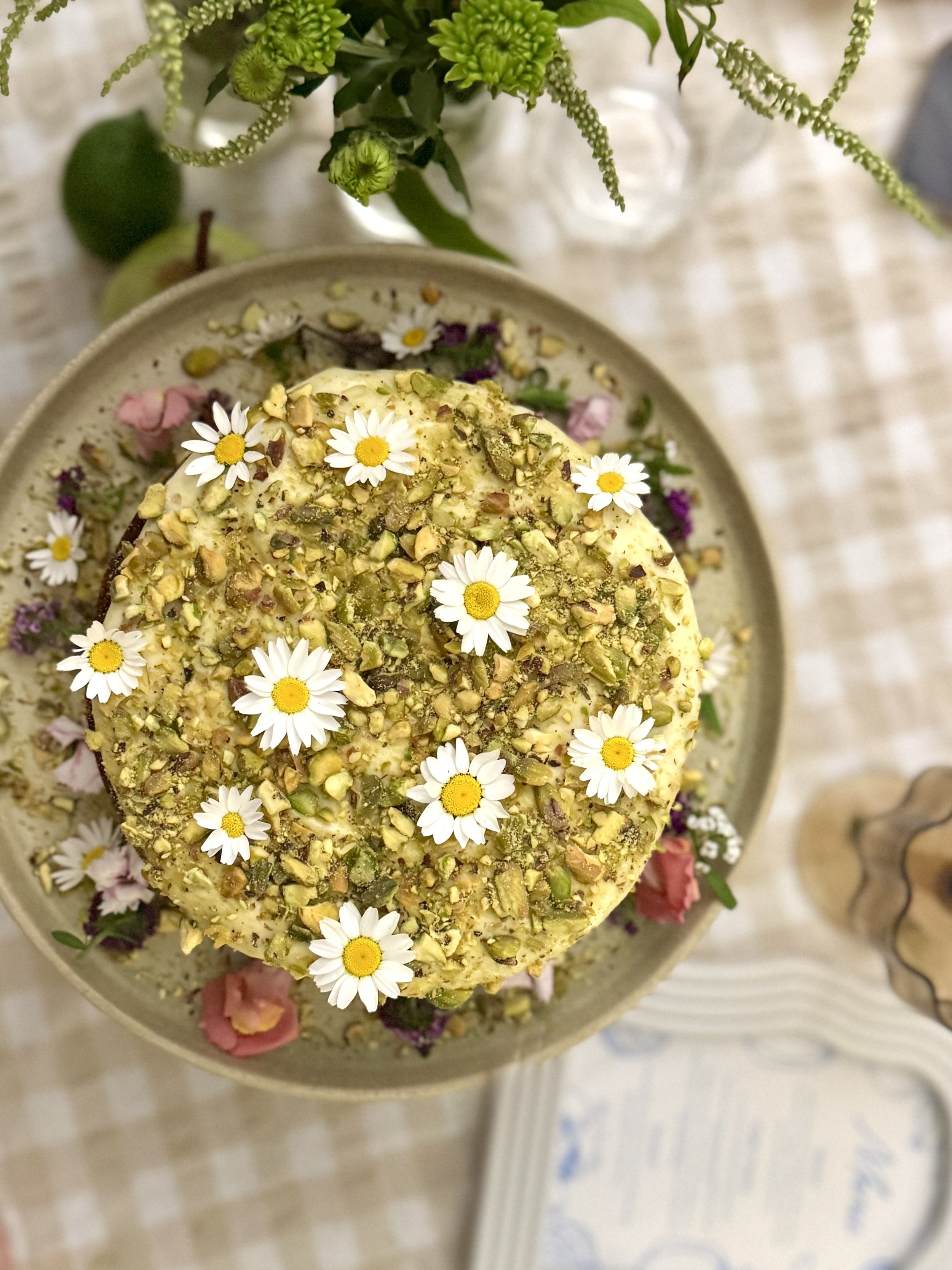 A cake topped with chopped pistachios and decorated with white daisy flowers, surrounded by various flowers and greenery on a table.
