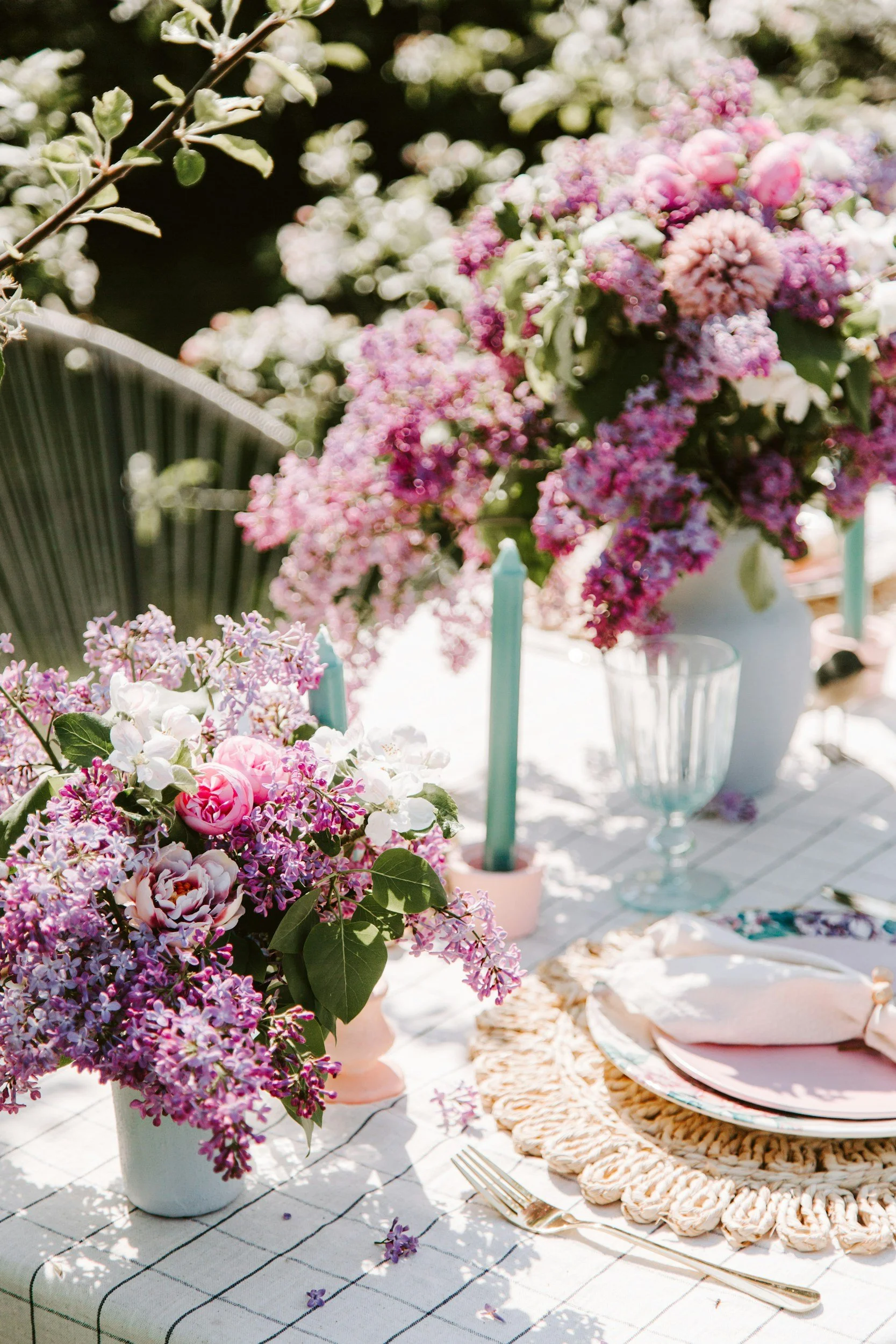 Decorated outdoor table with floral arrangements, pink and purple flowers, candles, glassware, and cutlery, set for a celebration or gathering.