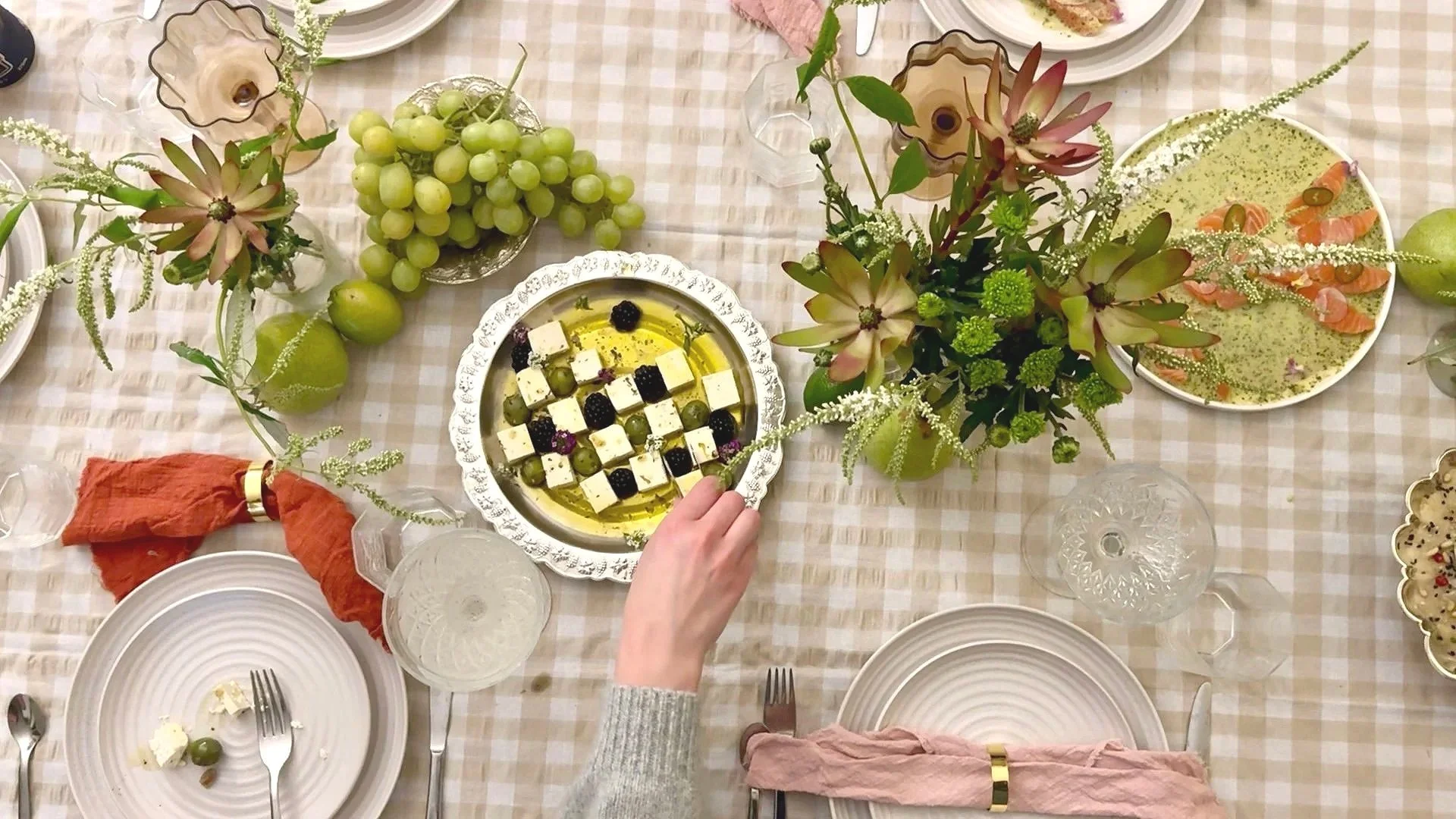 Top view of a table set for a meal with plates, glasses, cutlery, a centerpiece of flowers, and various dishes including cheese cubes with olives, grapes, and other appetizers.