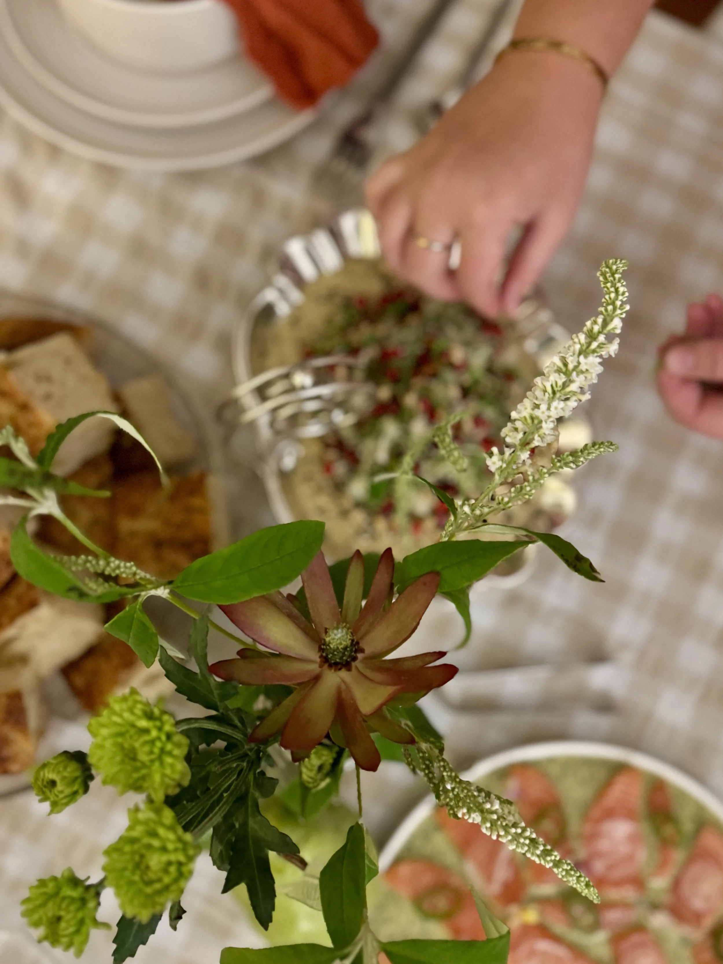 A person's hand reaching for a bowl of dip on a table set with plates, cutlery, bread, and a floral centerpiece.