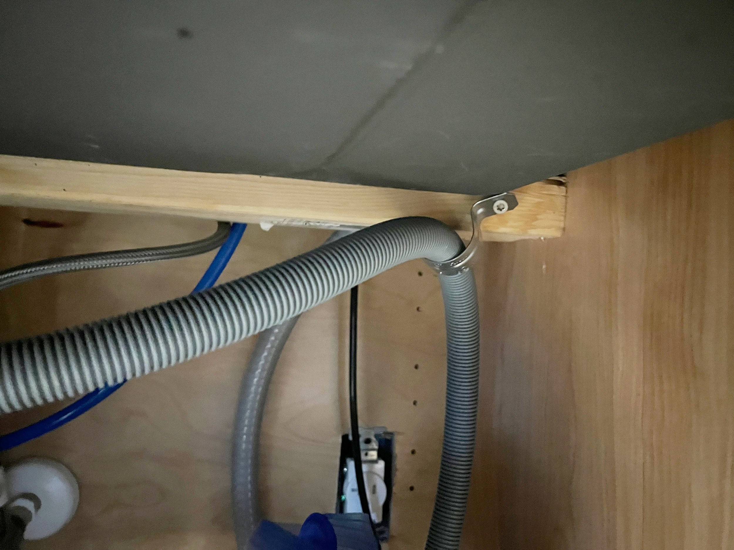 Under a kitchen or utility sink, there are several flexible hoses, including a silver corrugated drain hose, a gray hose, a blue water supply line, and a black cable. The hoses are attached to the cabinet's interior and the back of the sink.