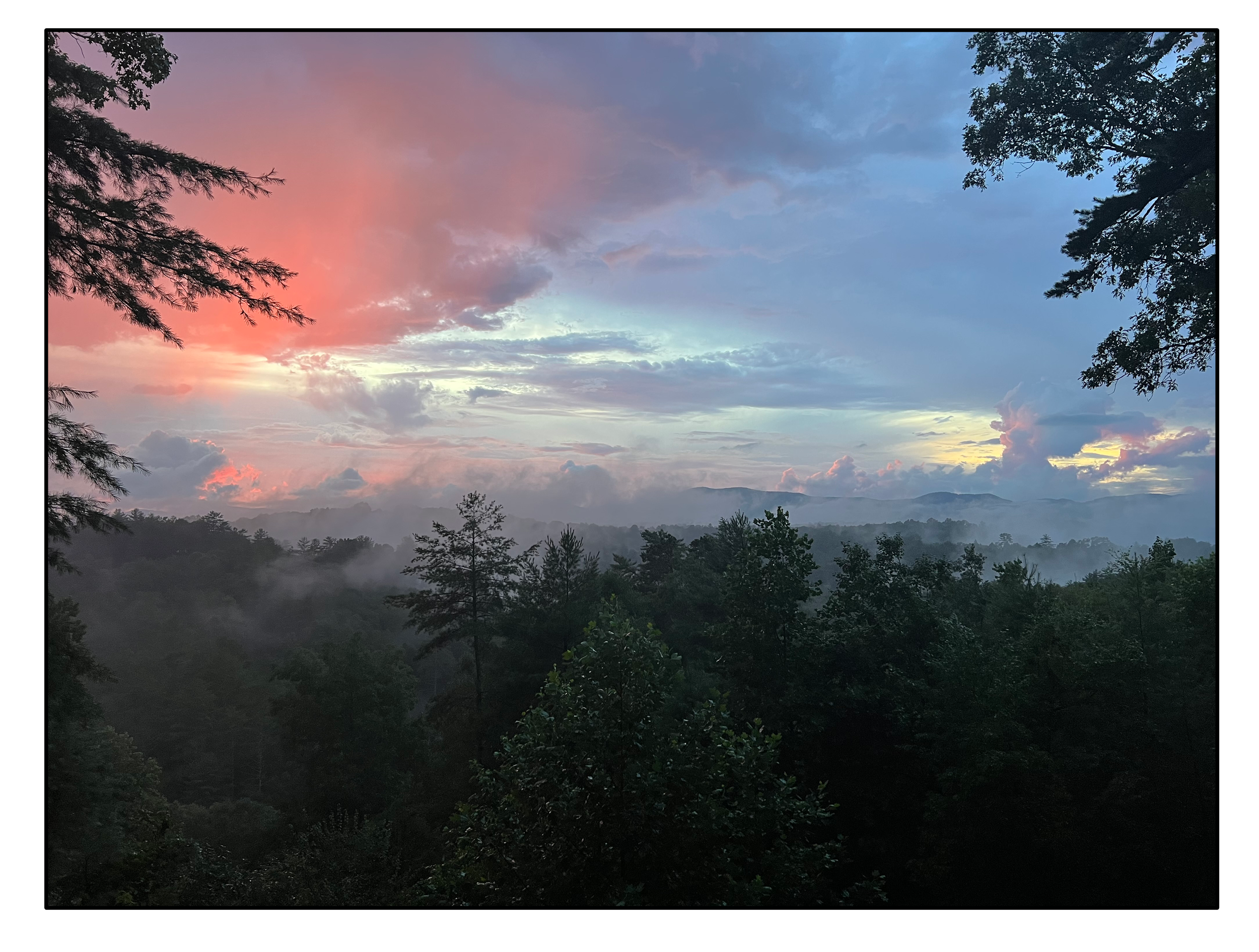 Scenic view of a forest at sunrise or sunset with colorful sky, clouds, and mountain silhouettes in the distance.