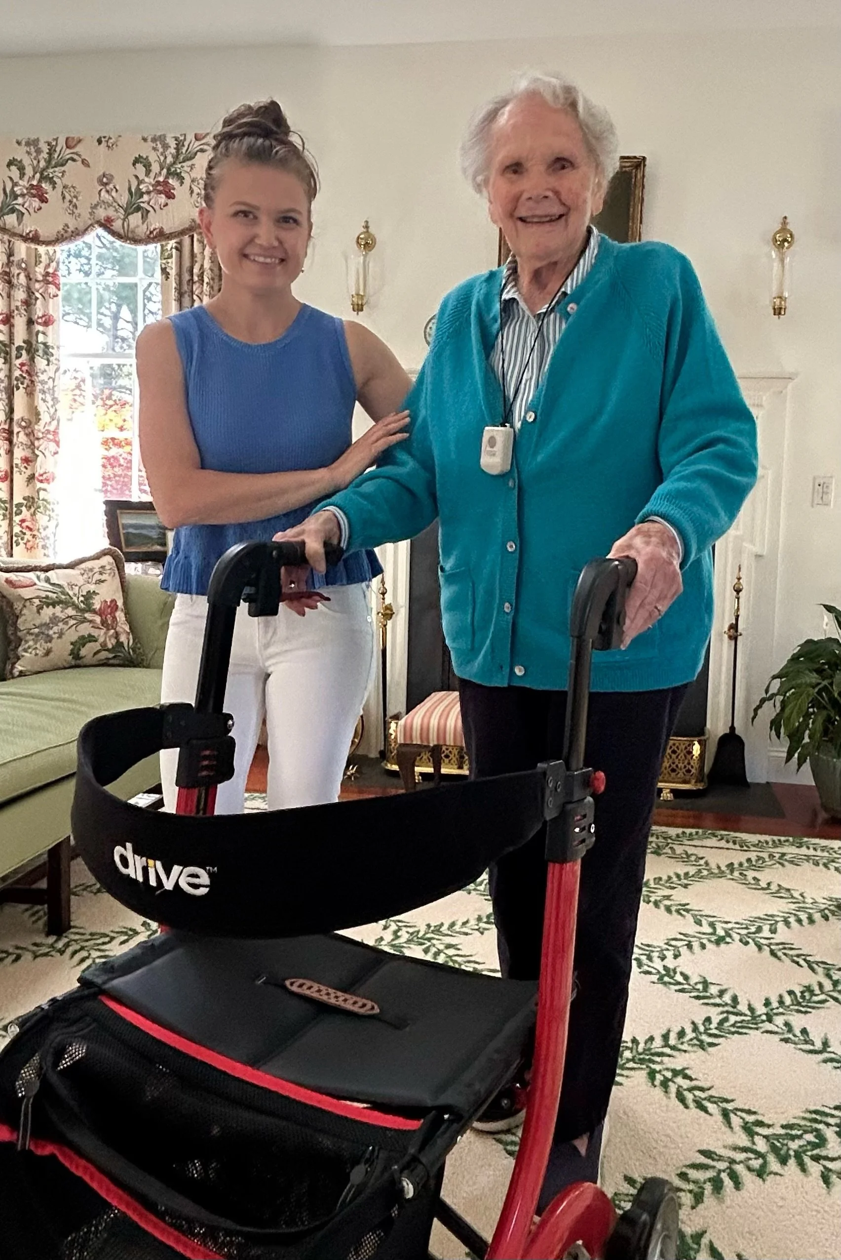 An elderly woman with white hair, using a walker, smiling, standing next to a younger woman with brown hair in a bun, wearing a blue sleeveless top and white pants, in a well-lit living room with floral curtains and decorated fireplace.
