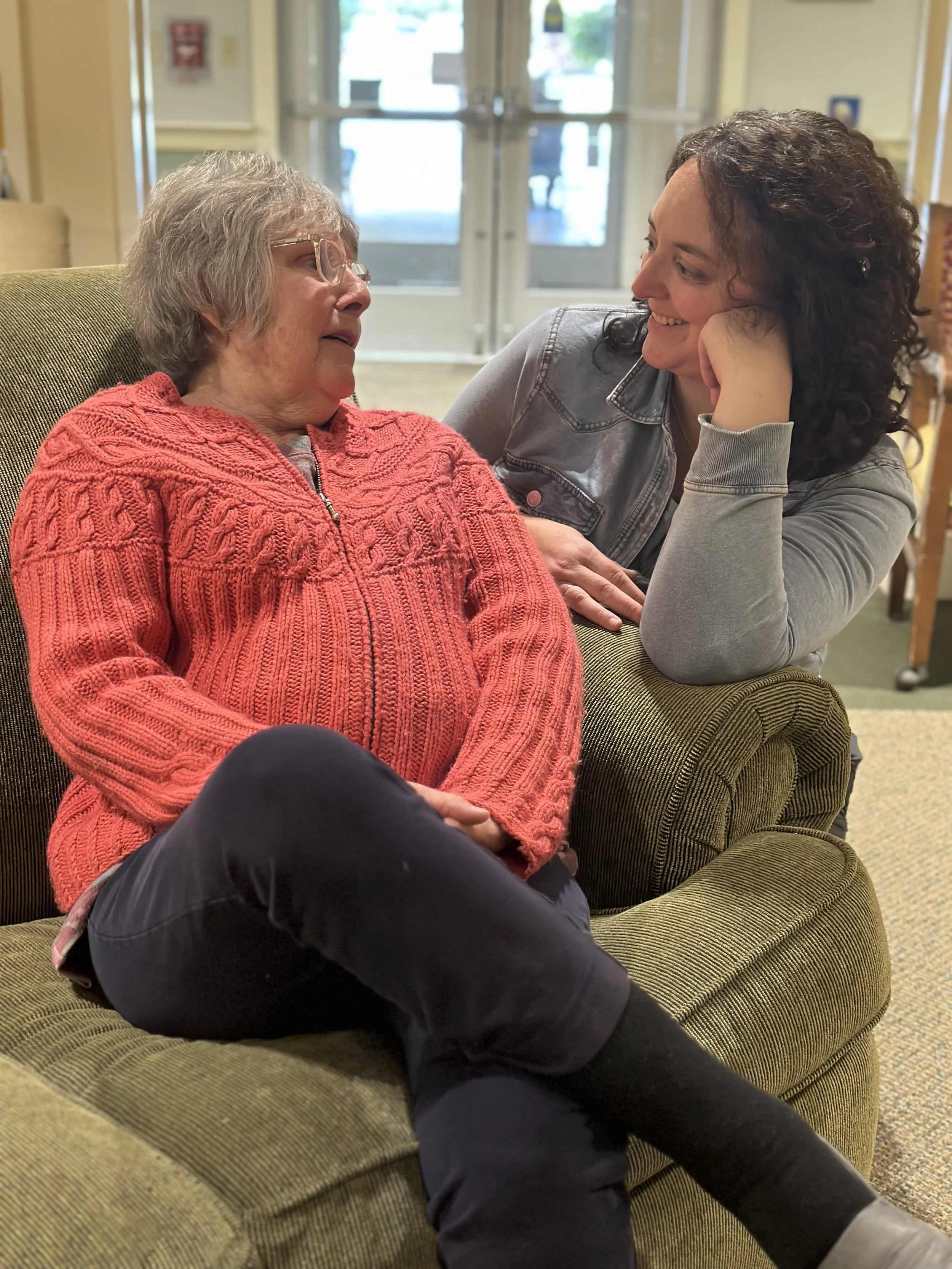 An elderly woman and a younger woman are sitting and leaning close to each other, engaged in a warm conversation inside a building, possibly a care facility or community center.