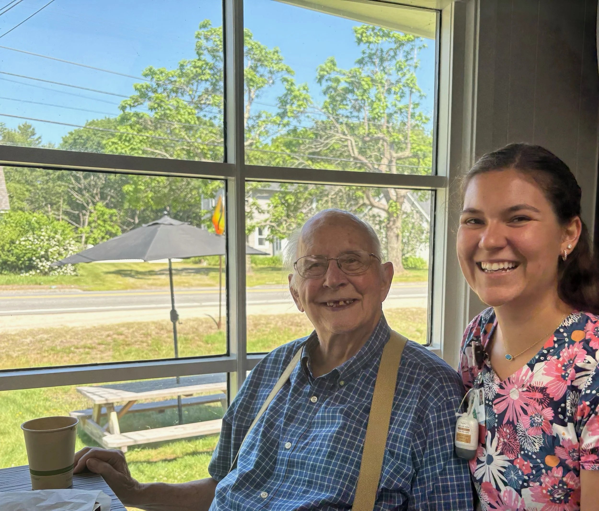A smiling elderly man with glasses and a young woman with dark hair, both sitting indoors near a window with a view of green trees, a picnic table, and a patio umbrella outside.