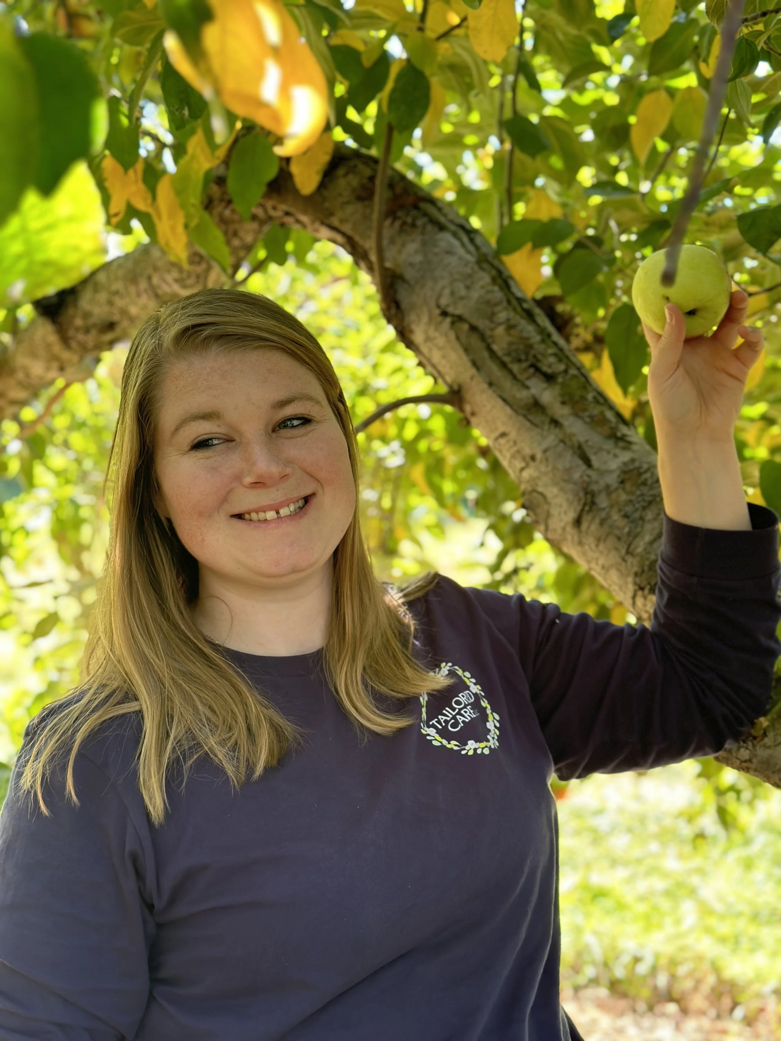A woman with long blonde hair smiling, holding a green apple on a tree branch surrounded by green and yellow leaves.