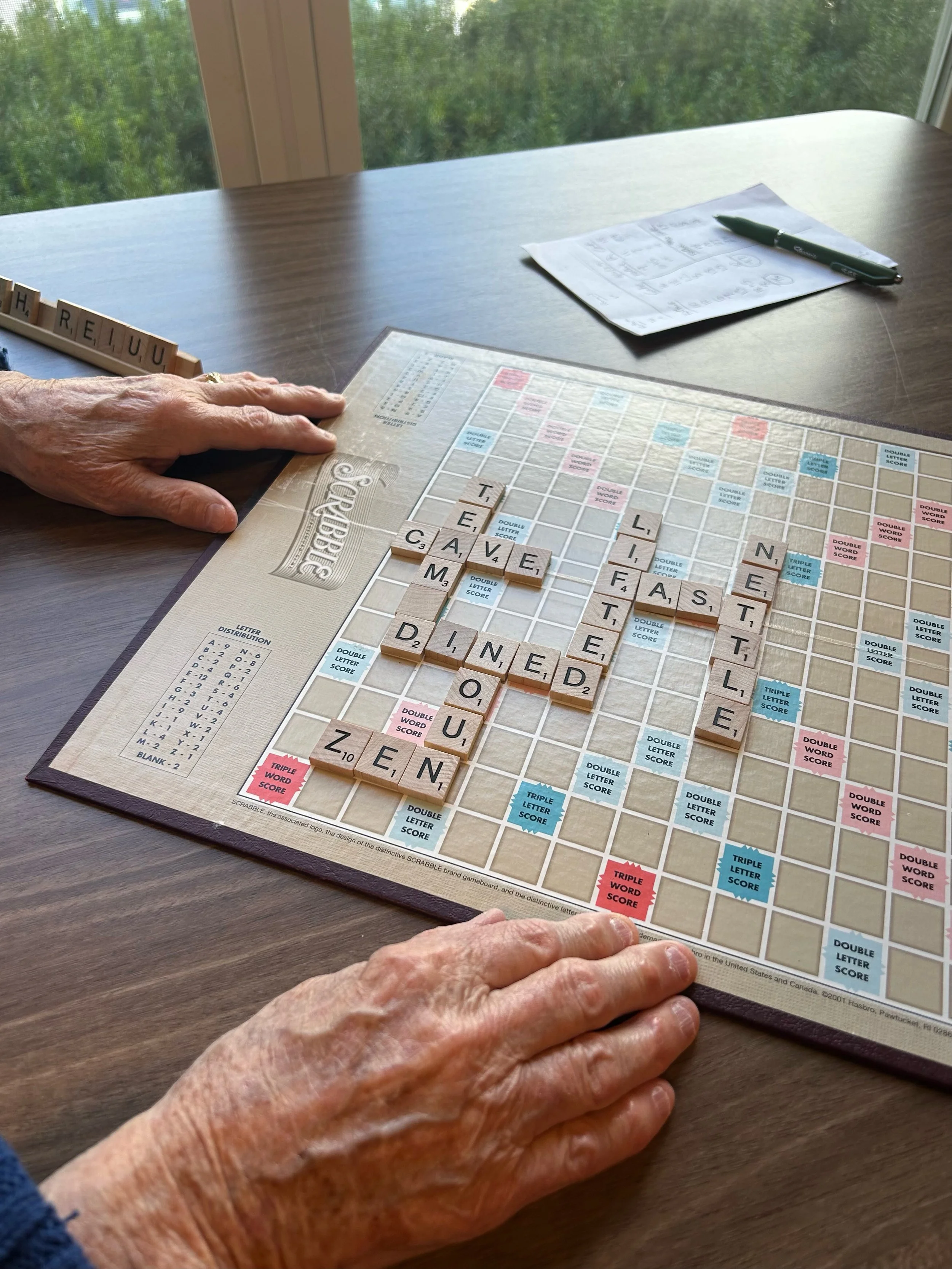 A person with elderly hands playing Scrabble at a wooden table near a window with greenery outside. The board displays words like 'CAVE', 'EAST', 'LINE', 'ZINE', and 'DONE'. A hand is holding Scrabble tiles spelling 'HREI JU'. There is a sheet of paper with notes and a green pen on the table.