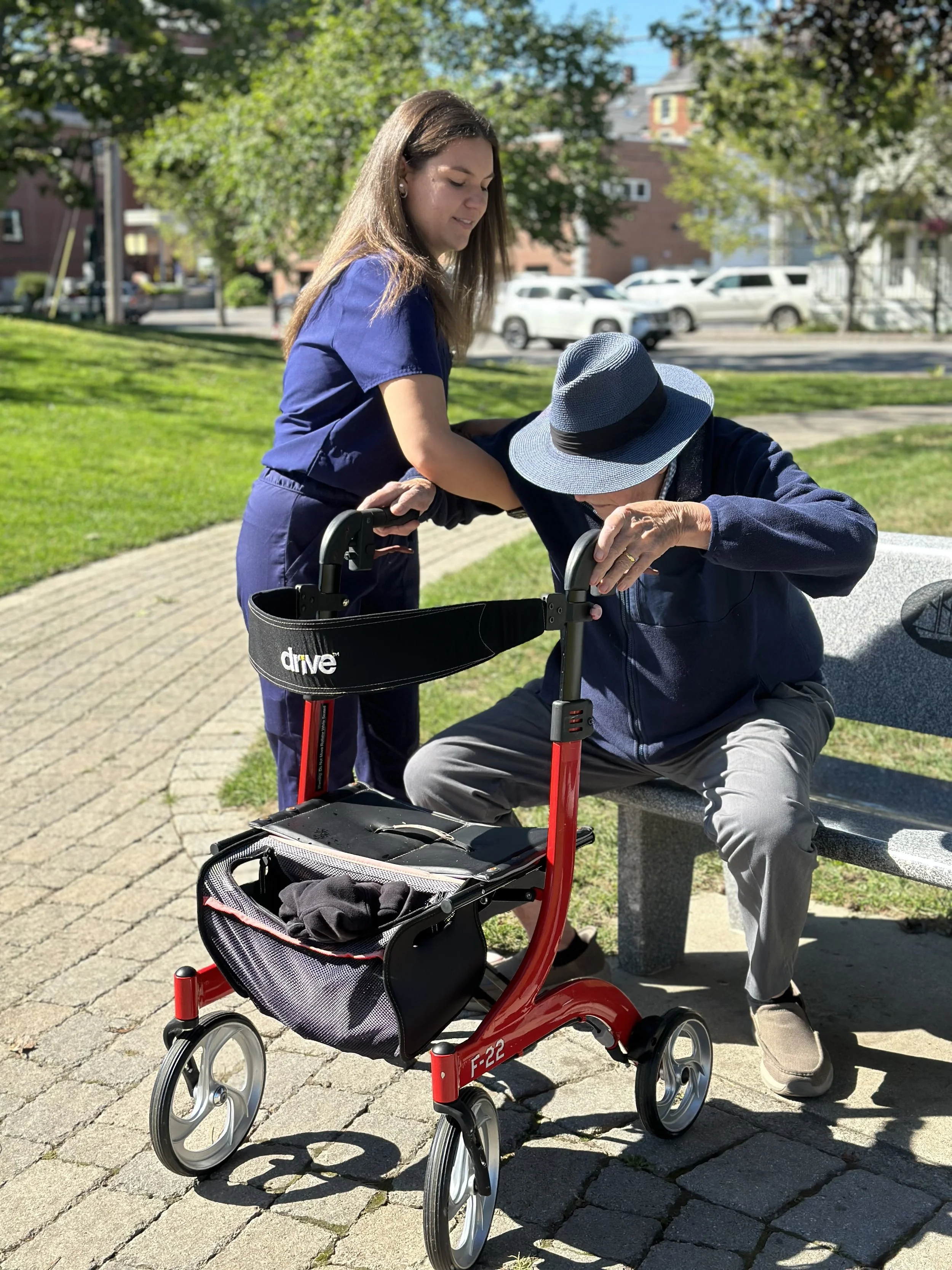 A young woman assisting an elderly man in a blue hat with a walker in a park on a sunny day.
