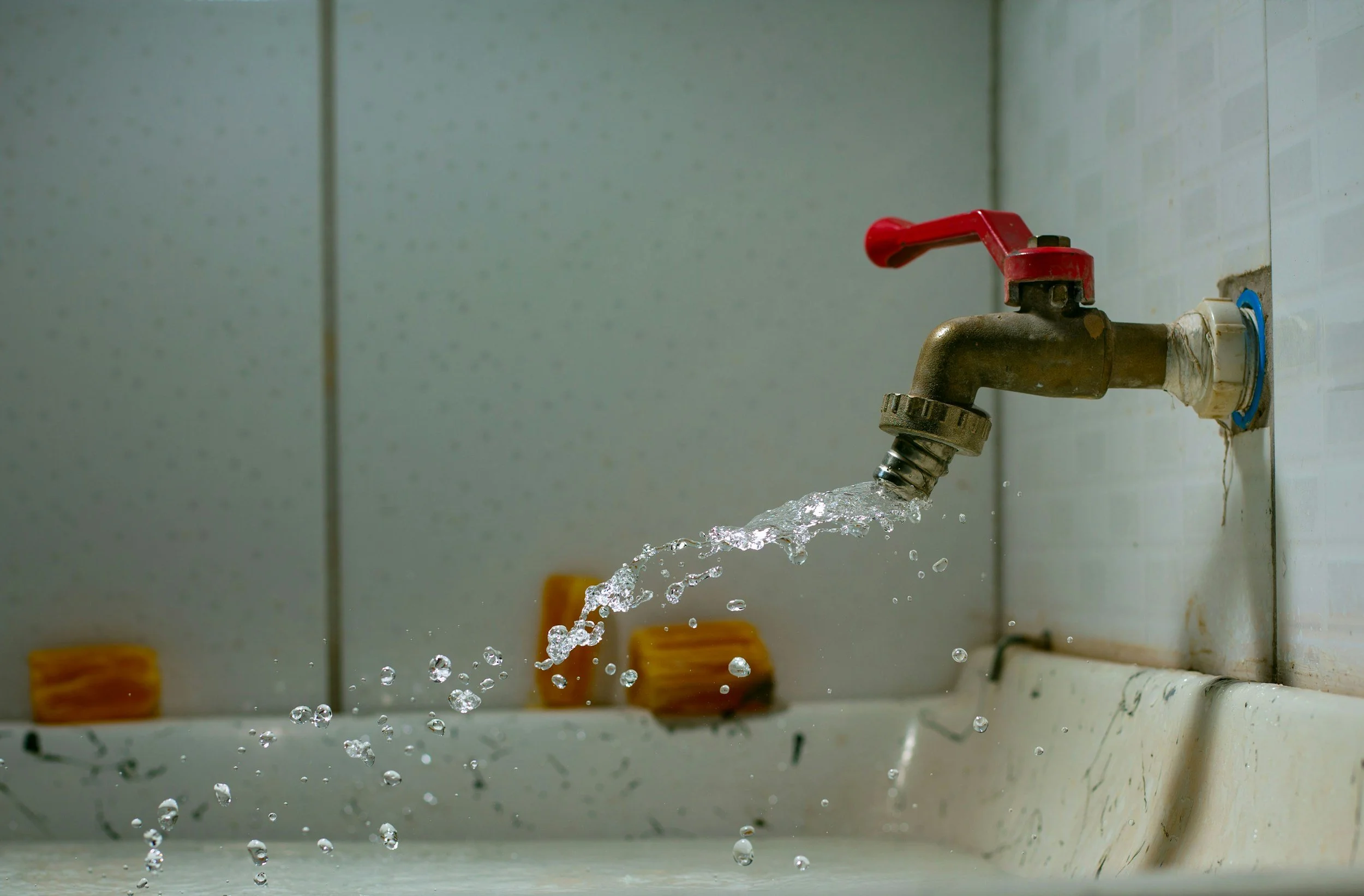 Un robinet en métal avec une poignée rouge, laissant couler de l'eau dans un évier en céramique, avec deux éponges en arrière-plan