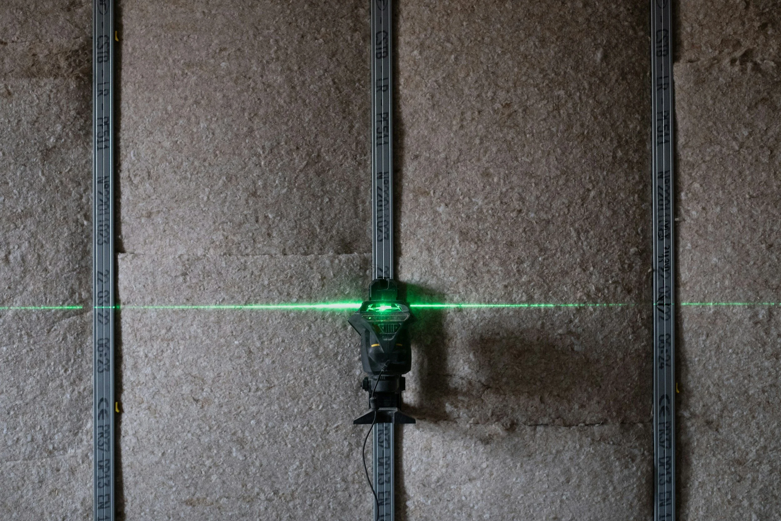 Un niveau laser vert fixé sur un mur en pierre, avec deux rails métalliques verticaux fixés en parallèle.