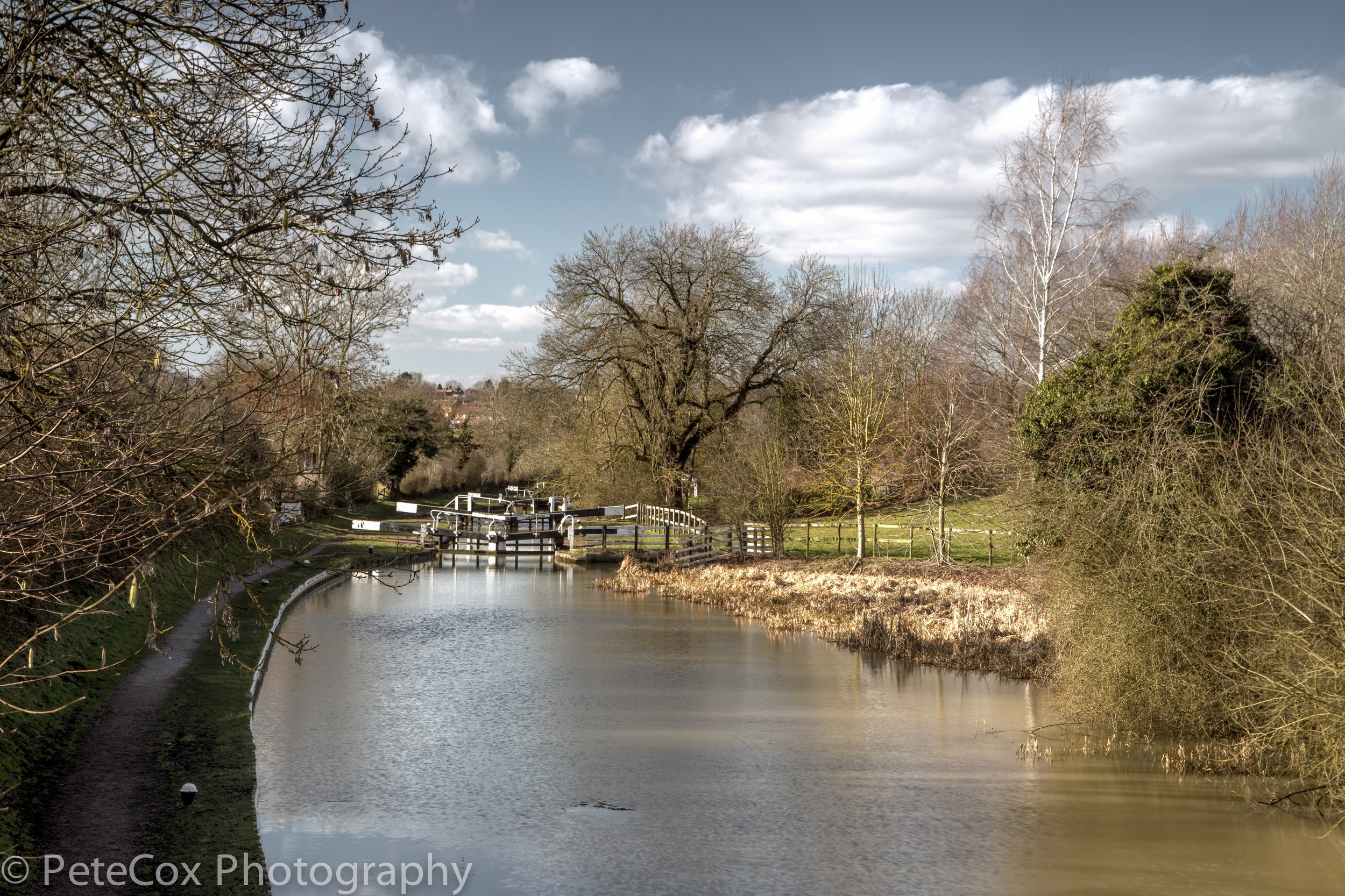 A river with a lock system in a rural landscape, surrounded by leafless trees and a grassy path on one side, with a blue sky and clouds overhead.