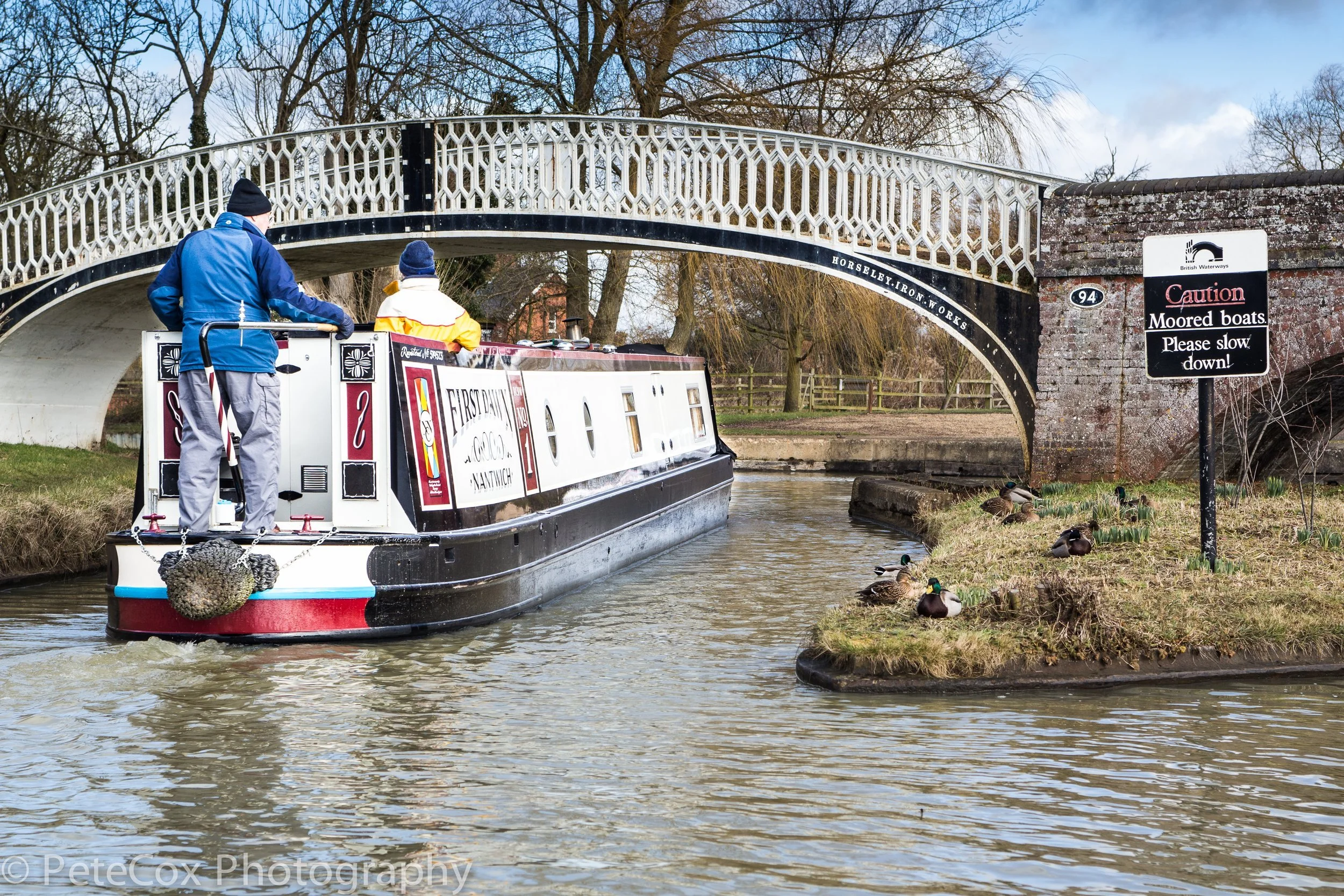 A narrowboat navigating a canal underneath a decorative iron footbridge, with ducks on the grassy bank nearby. A sign warns about moored boats and advises to slow down.