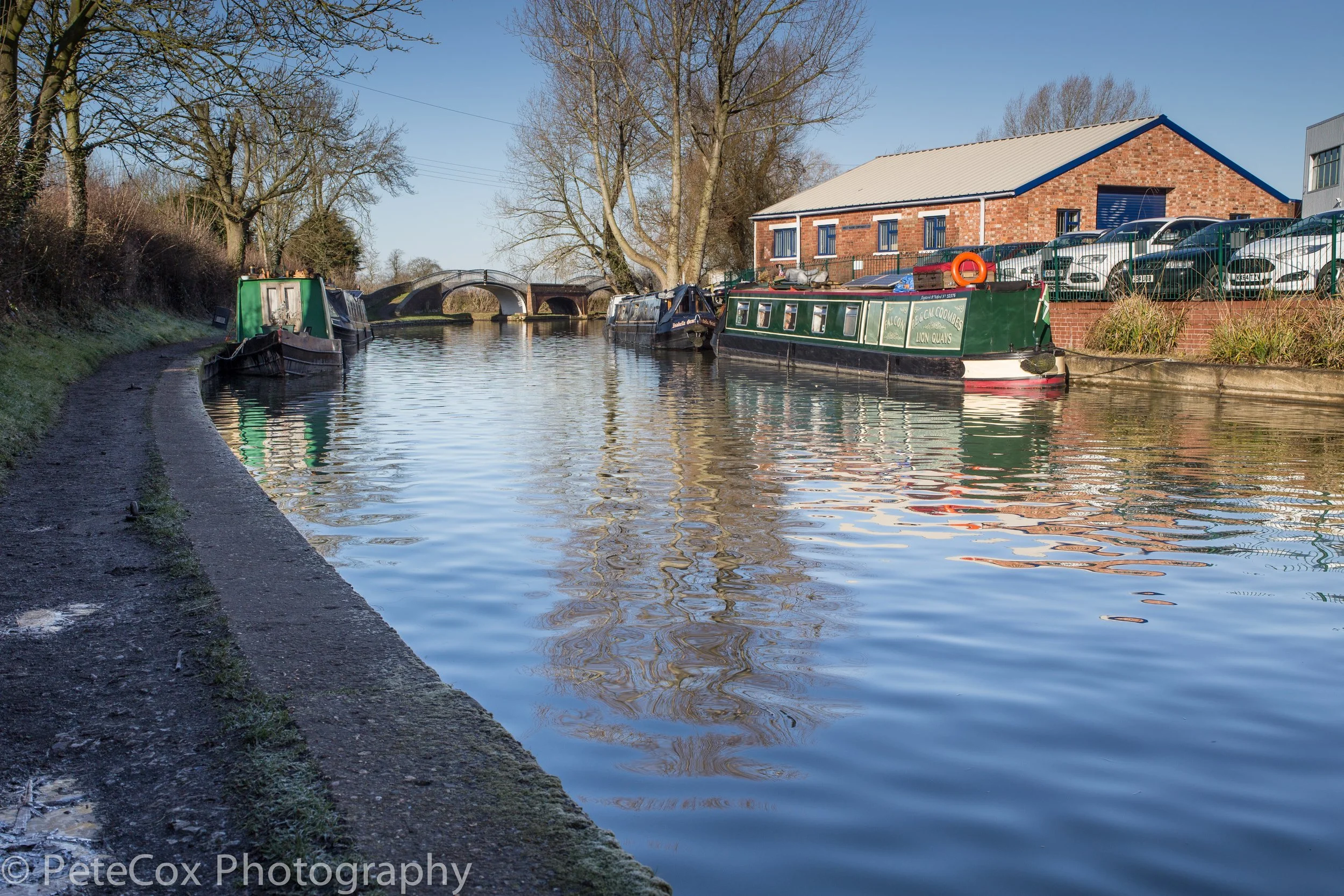 A canal scene with multiple narrowboats moored along the right side, a stone bridge in the background, a brick building with parked cars on the right, and leafless trees lining the canal on the left, under a clear blue sky.