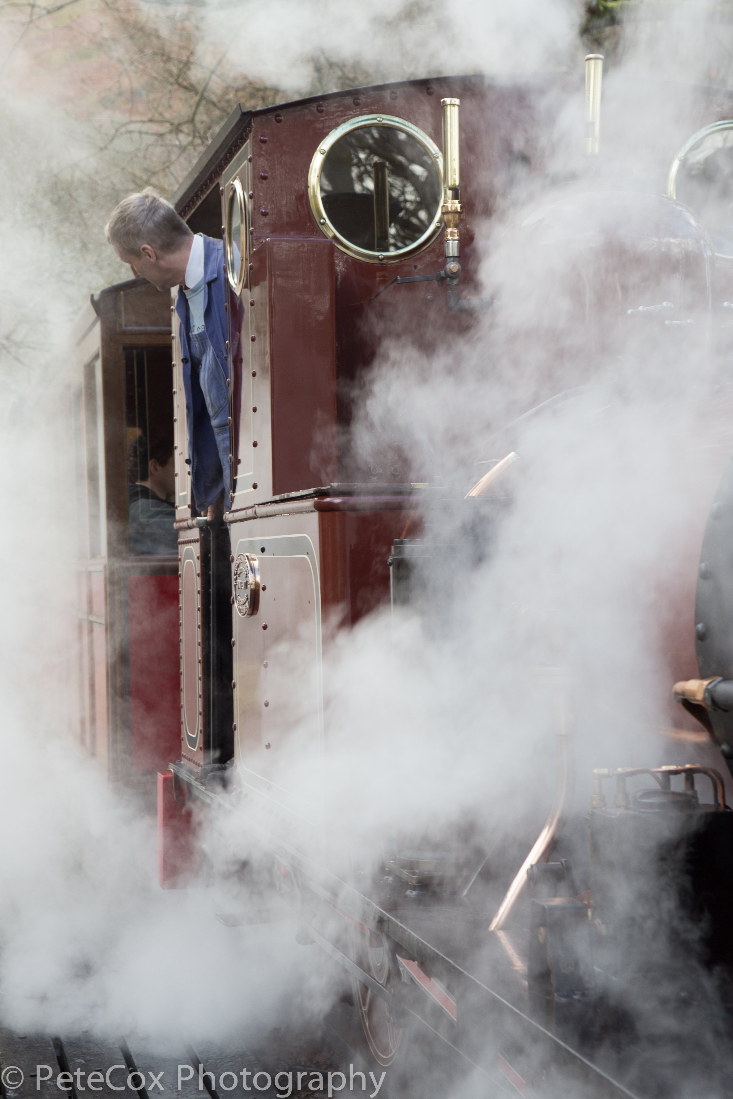 A vintage steam train emitting steam, with a man in a blue jacket leaning out of the window.