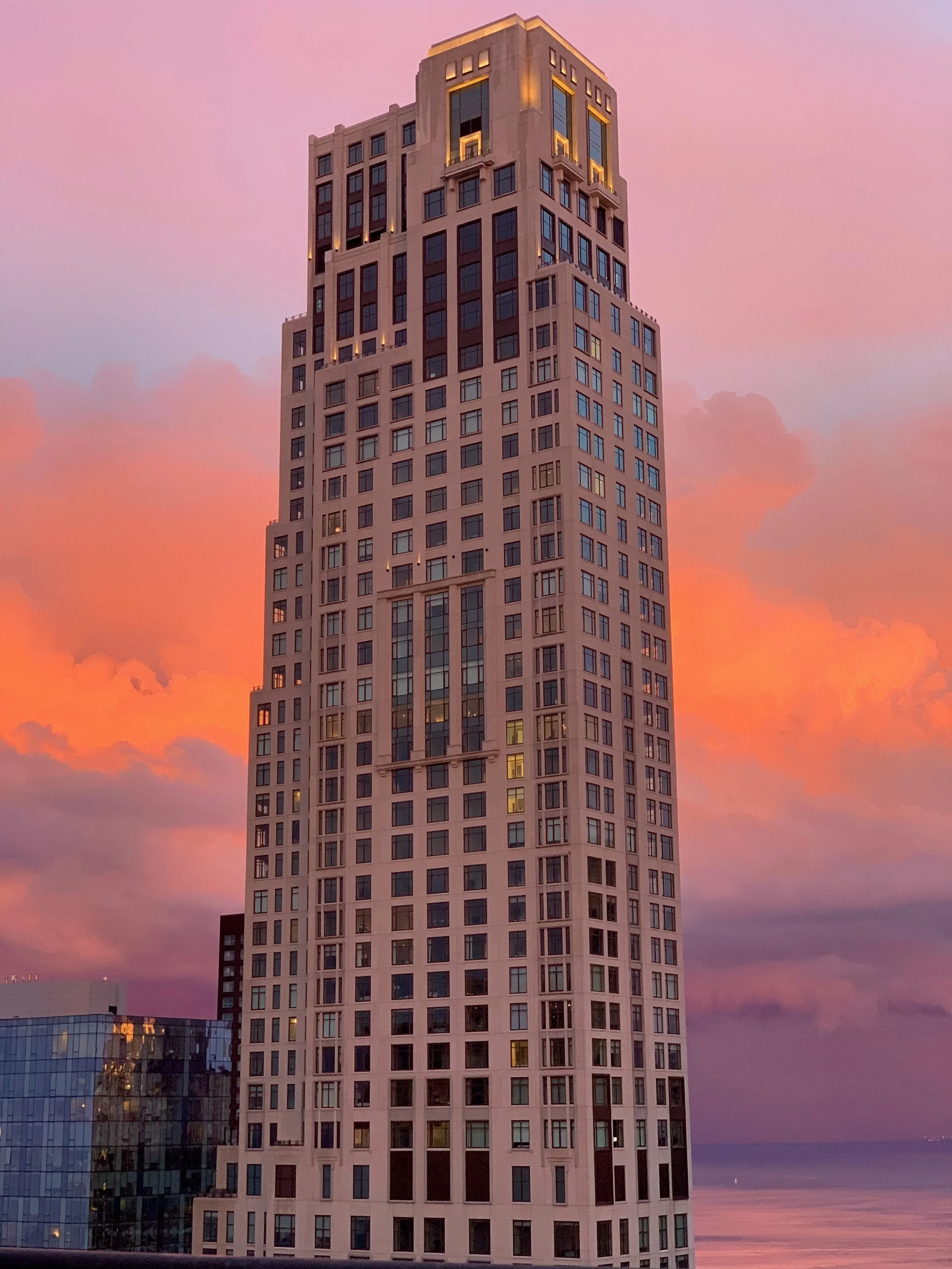A tall skyscraper with numerous windows, illuminated at the top, against a pink and purple sunset sky.