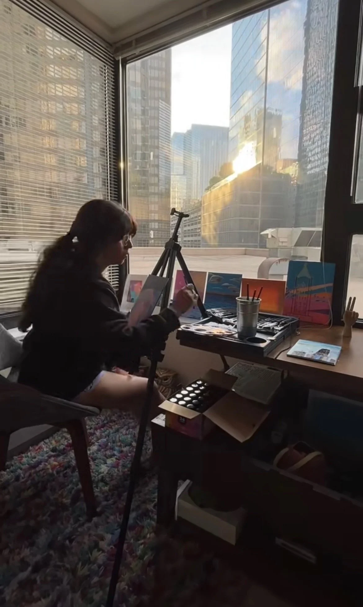 A young woman sitting by a window, painting on a canvas with art supplies and paintings around in an urban high-rise art studio.